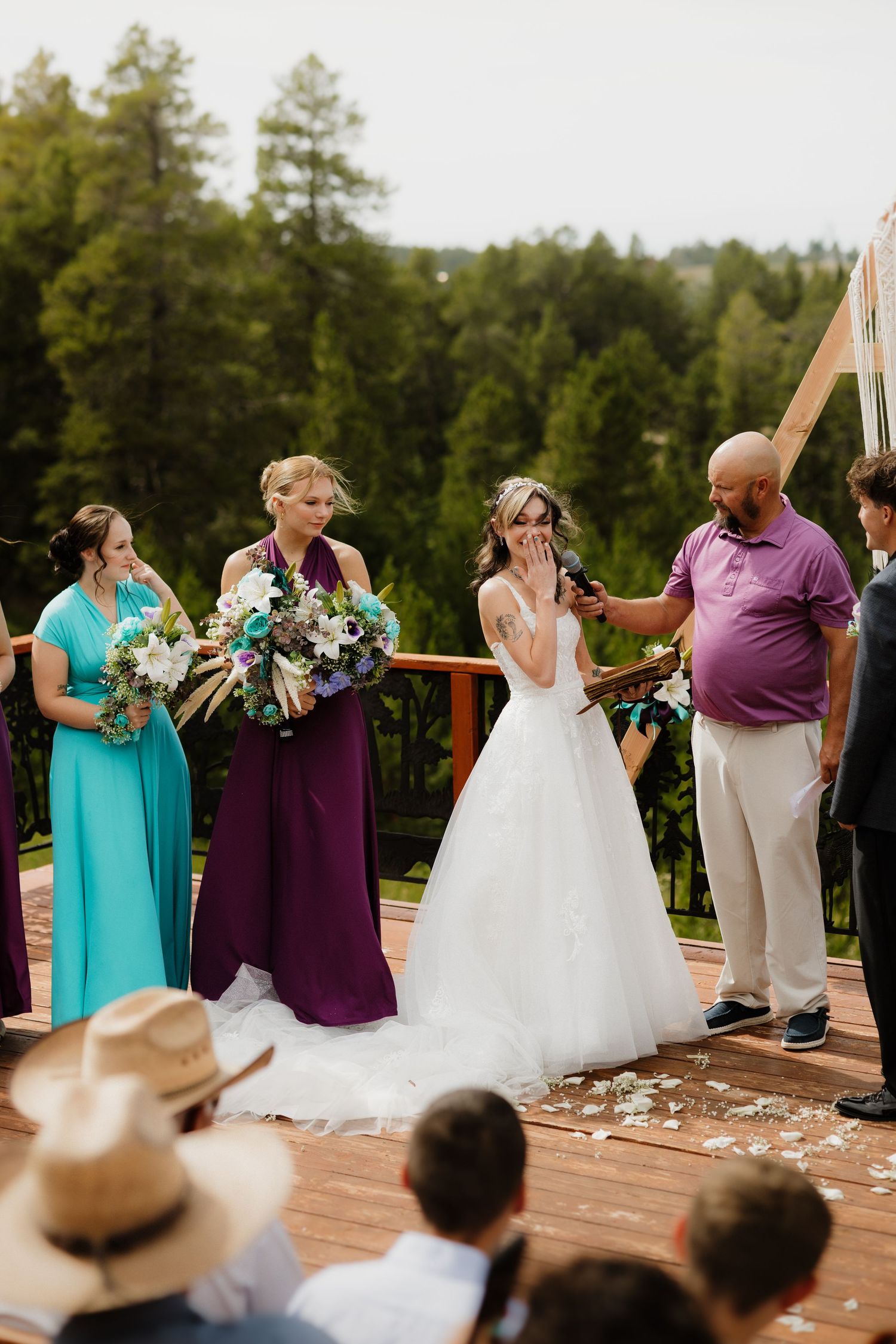 Emotional bride covering her mouth during outdoor wedding ceremony on a wooden deck with bridesmaids.