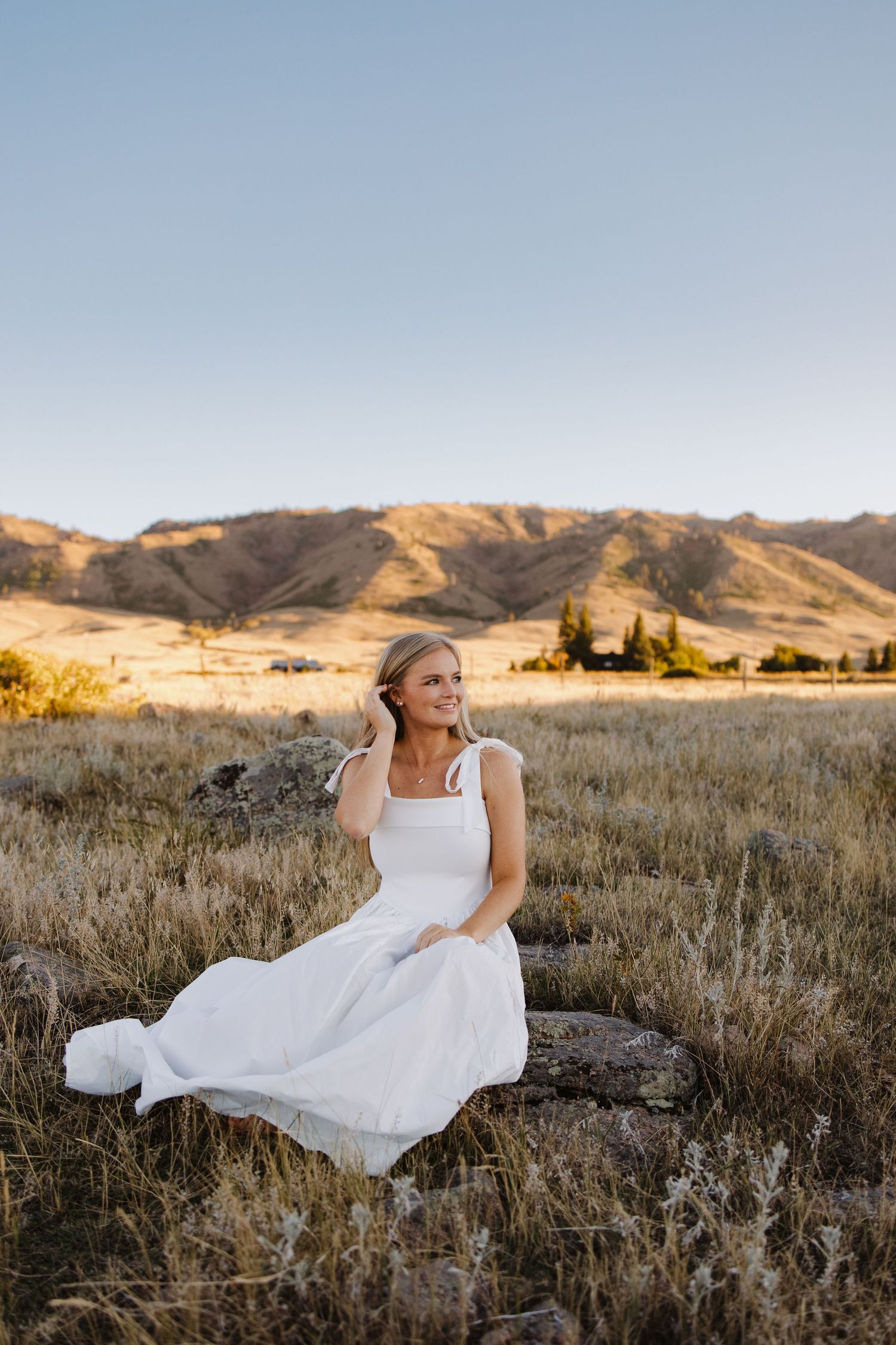 Smiling bride in white dress sits on rocks in golden field with mountain backdrop at sunset.