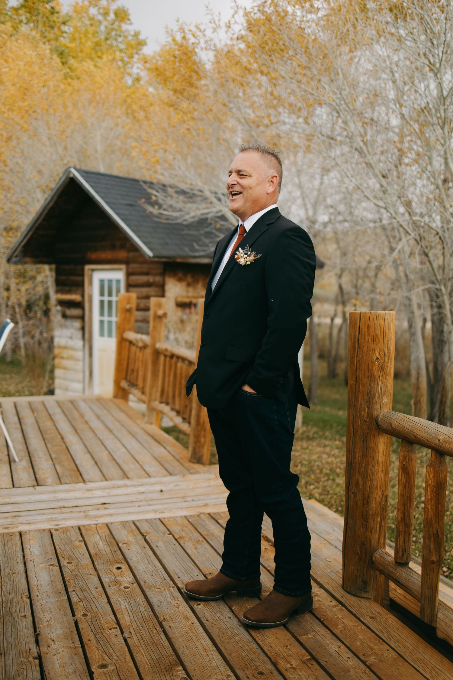 Smiling groom in black suit stands on wooden deck surrounded by autumn foliage.