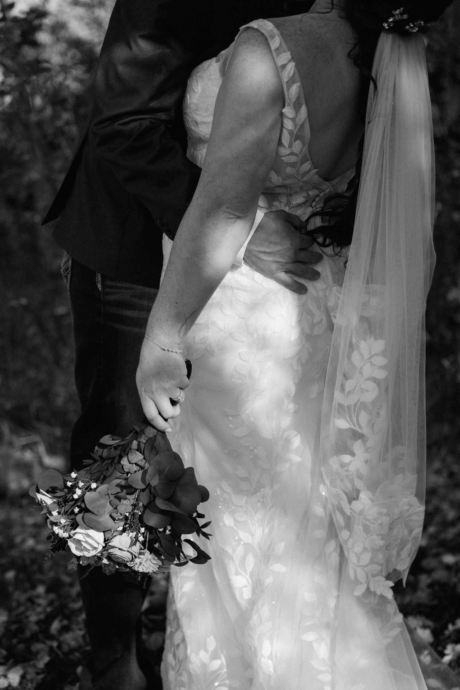 Black and white close-up of bride in lace gown holding bouquet with groom embracing her.
