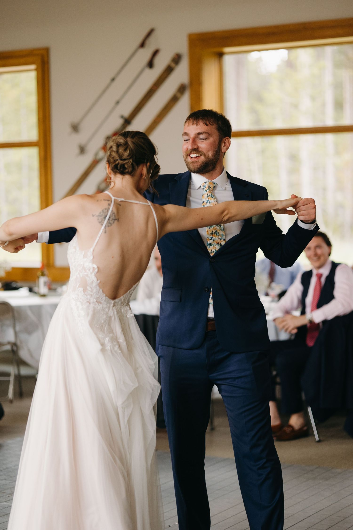 Smiling groom in navy suit shares first dance with bride in backless lace wedding gown indoors.