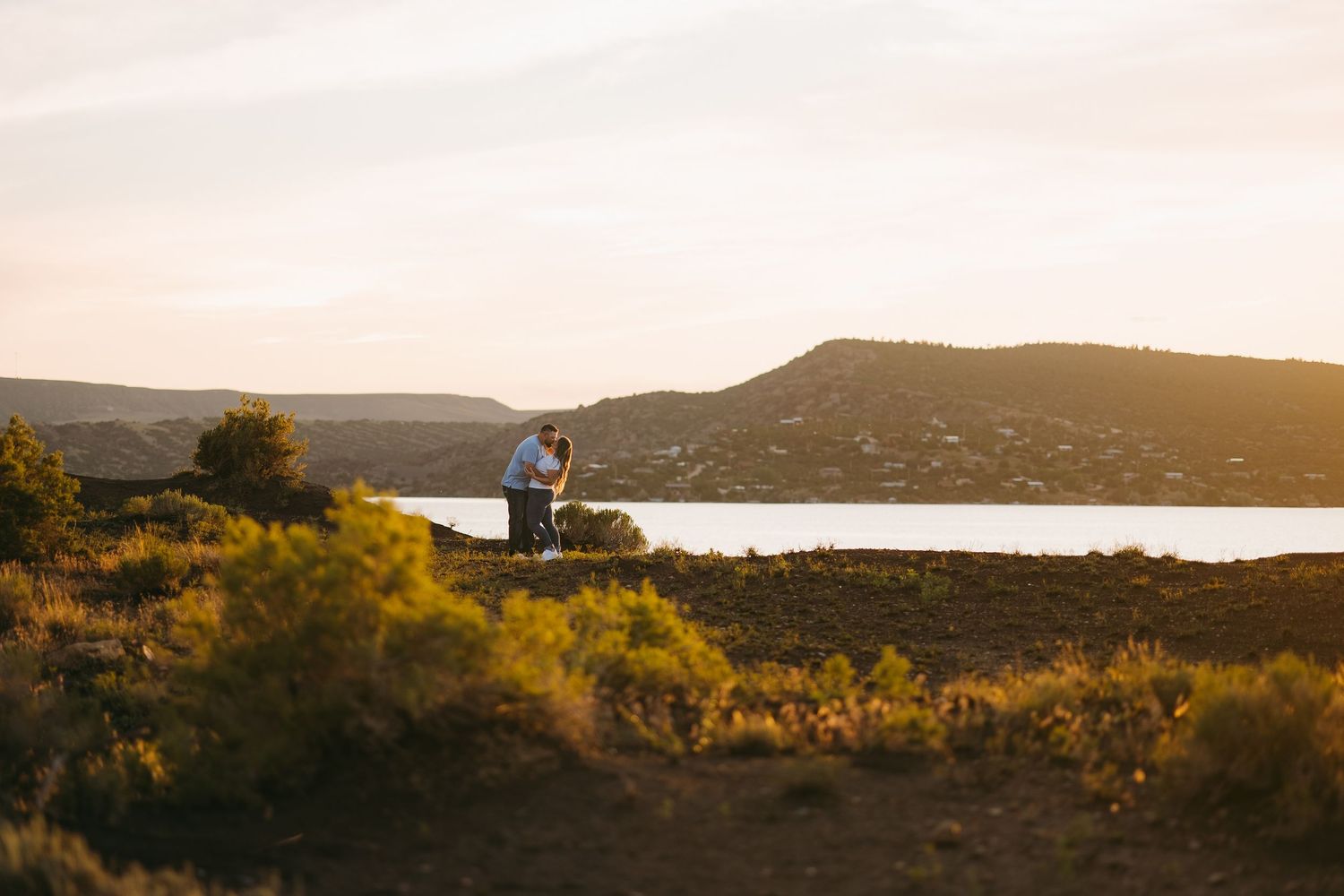 Couple embraces at golden hour on a scenic overlook above a shimmering lake and hillside.
