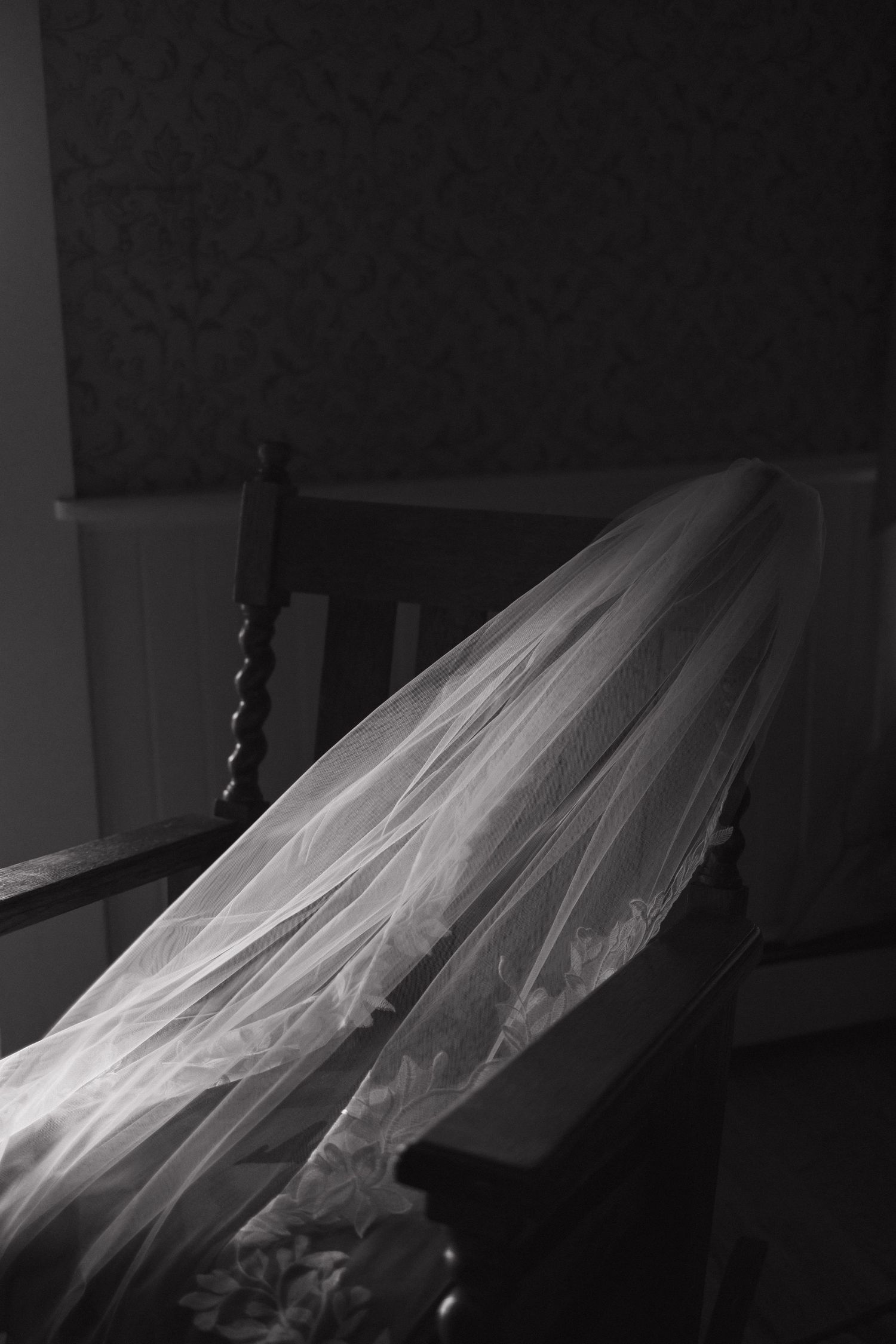 Delicate bridal veil draped over a wooden chair captured in soft black and white lighting.