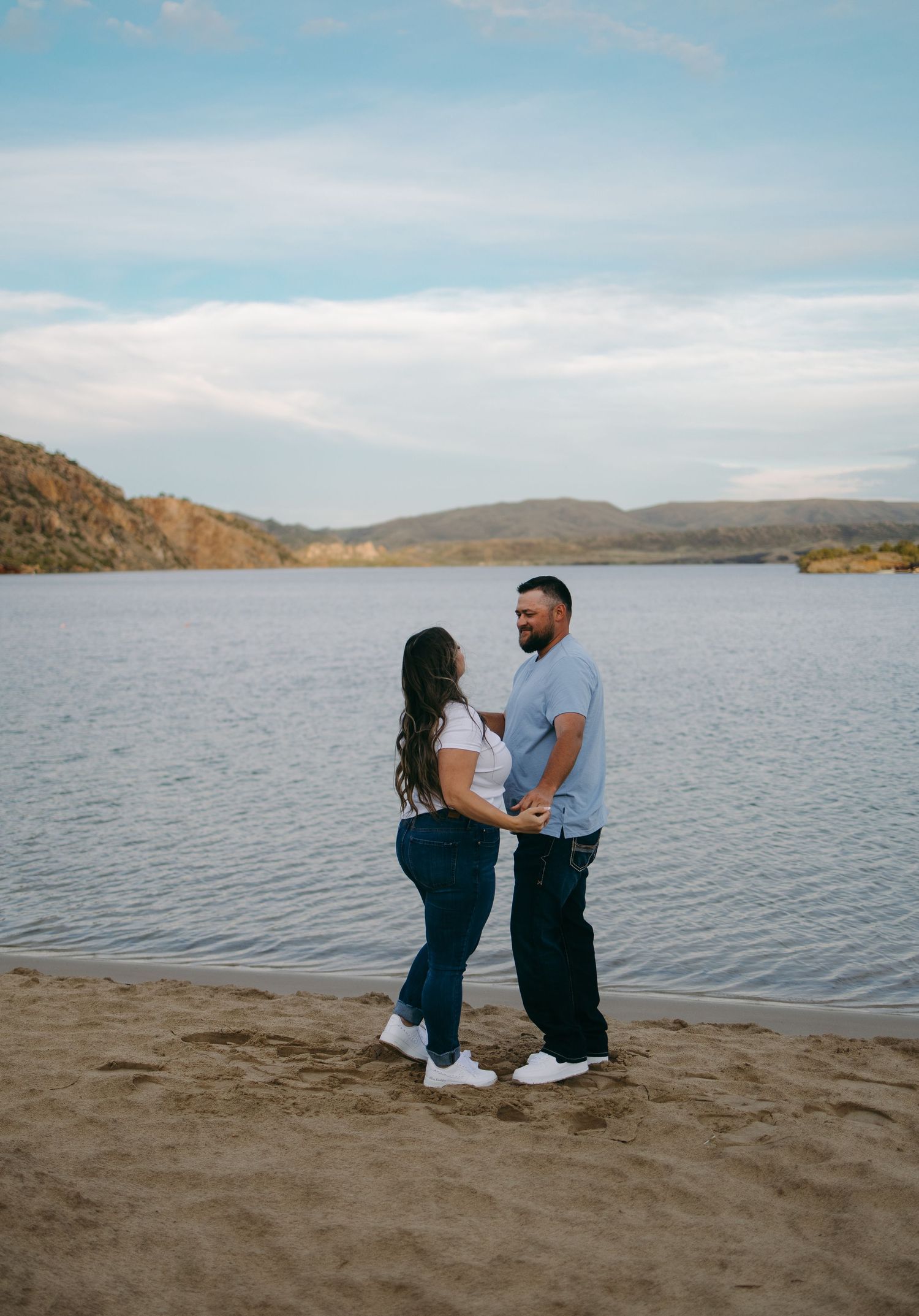Couple holds hands and smiles facing each other on a sandy lakeshore under a cloudy sky.