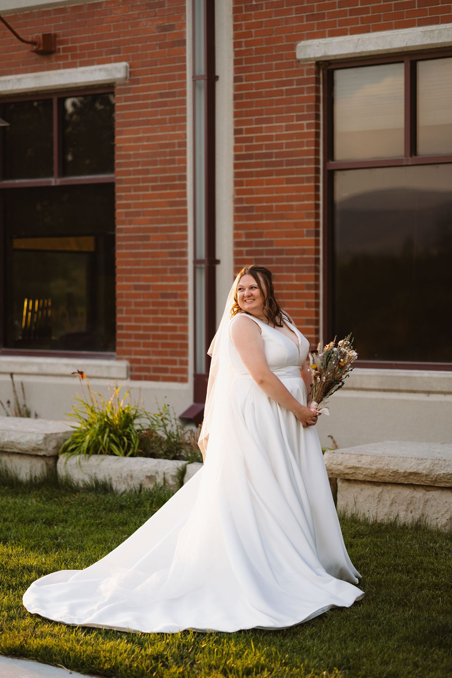 Bride in elegant white A-line gown with veil holds bouquet while posing in front of brick building.