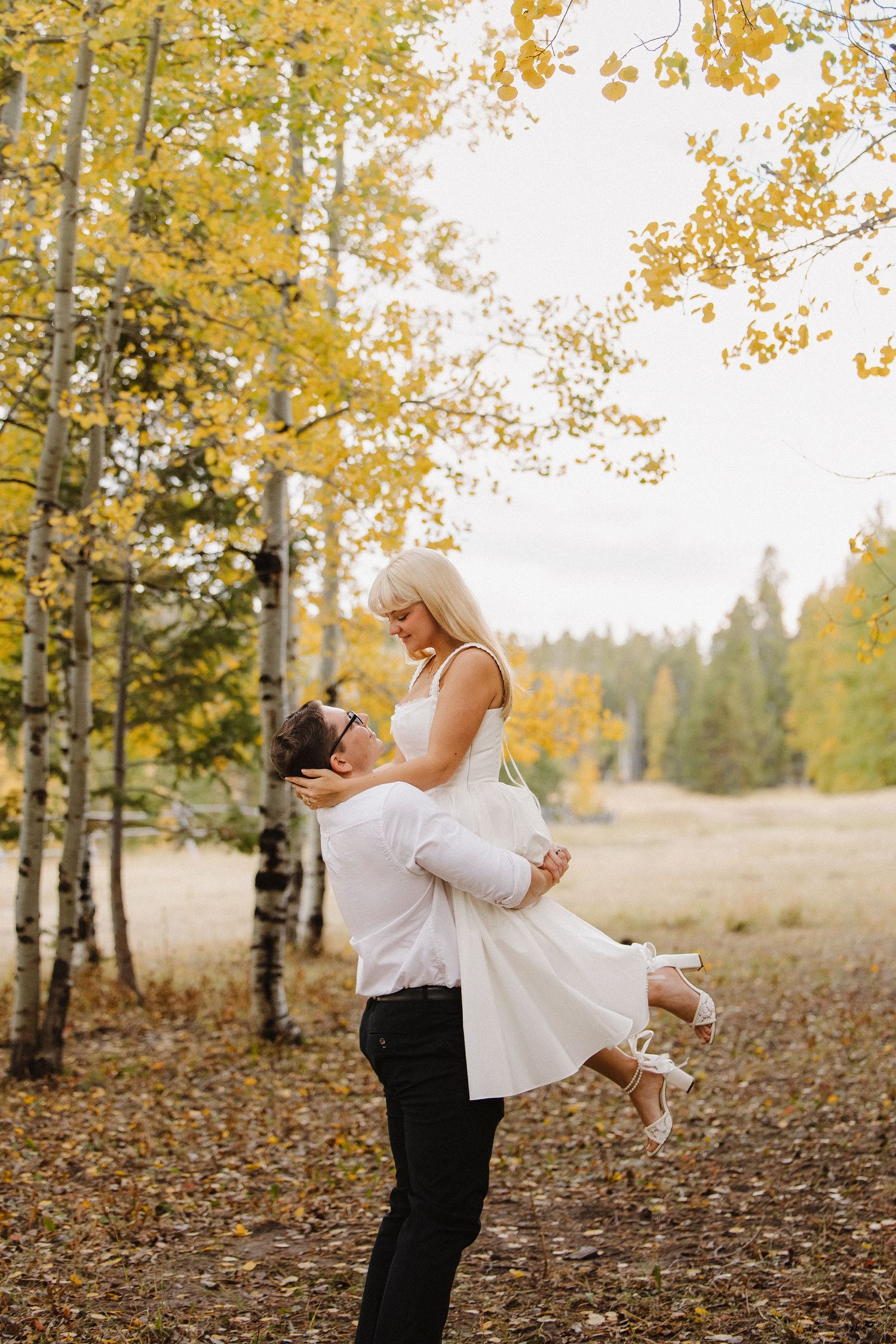 Groom lifts bride in short white dress beneath golden aspen trees during autumn wedding portraits. Wyoming wedding.