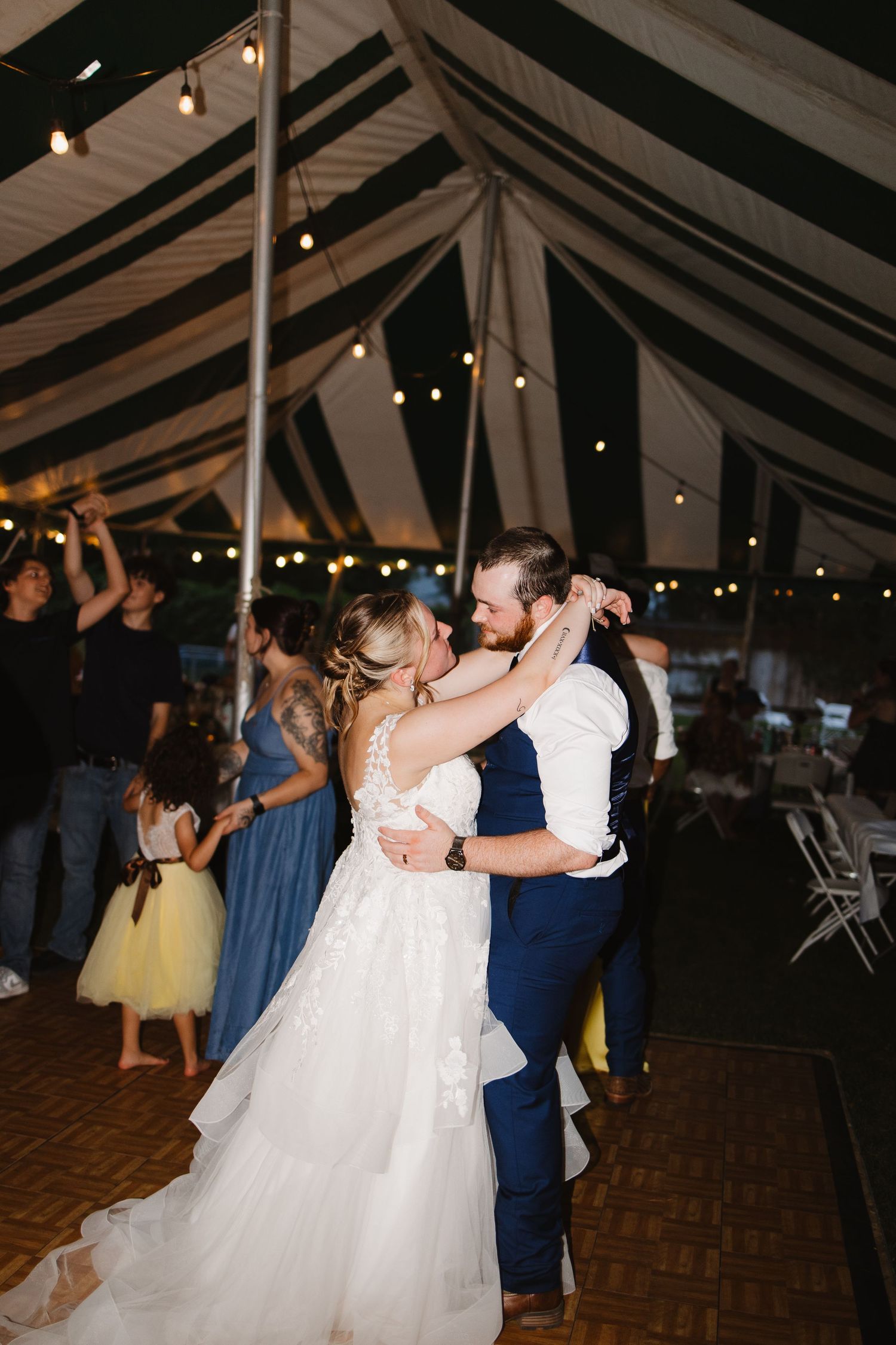 Bride and groom share first dance under string lights in a striped wedding tent surrounded by guests.