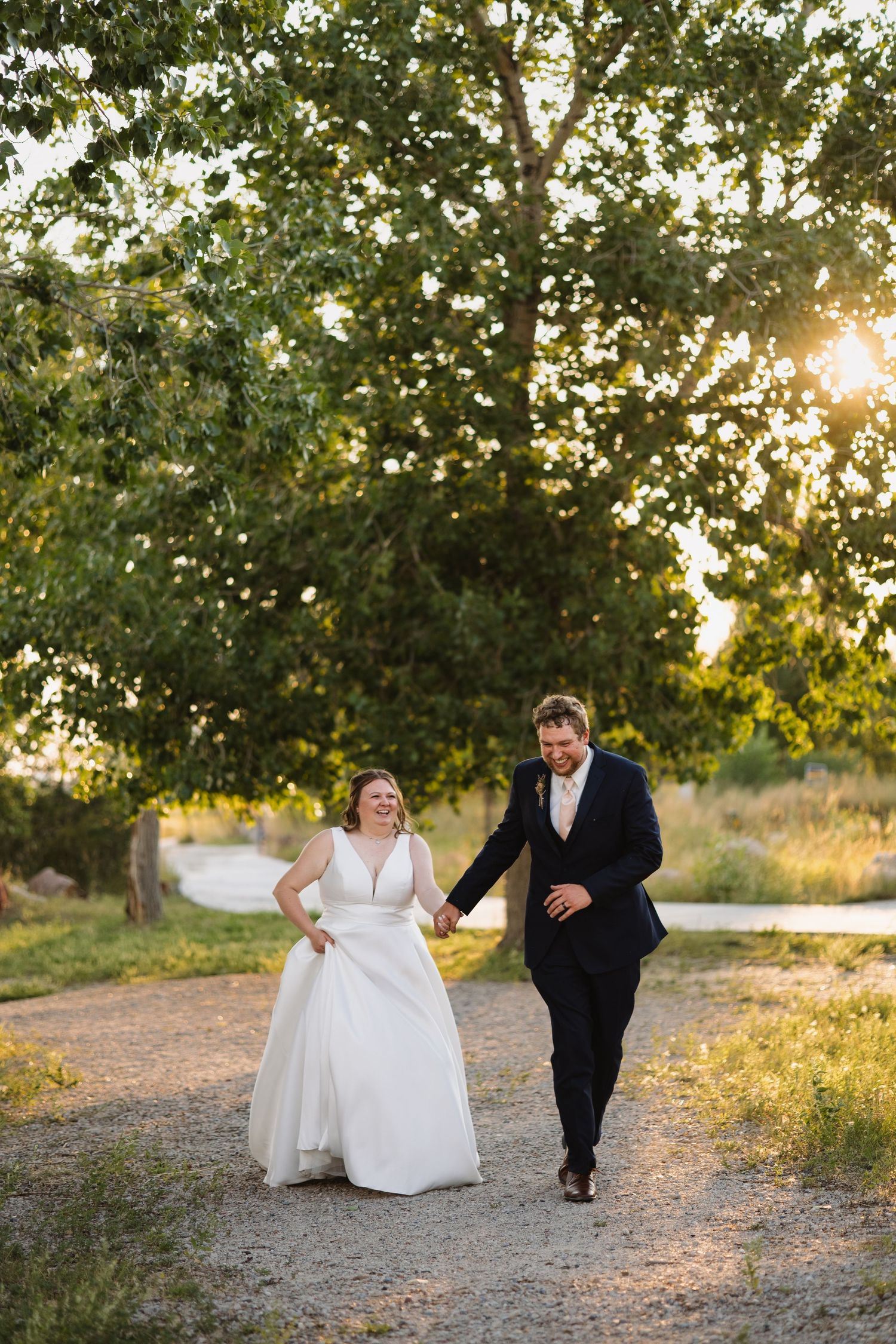 Laughing bride and groom walk hand in hand down a gravel path during golden hour portraits.