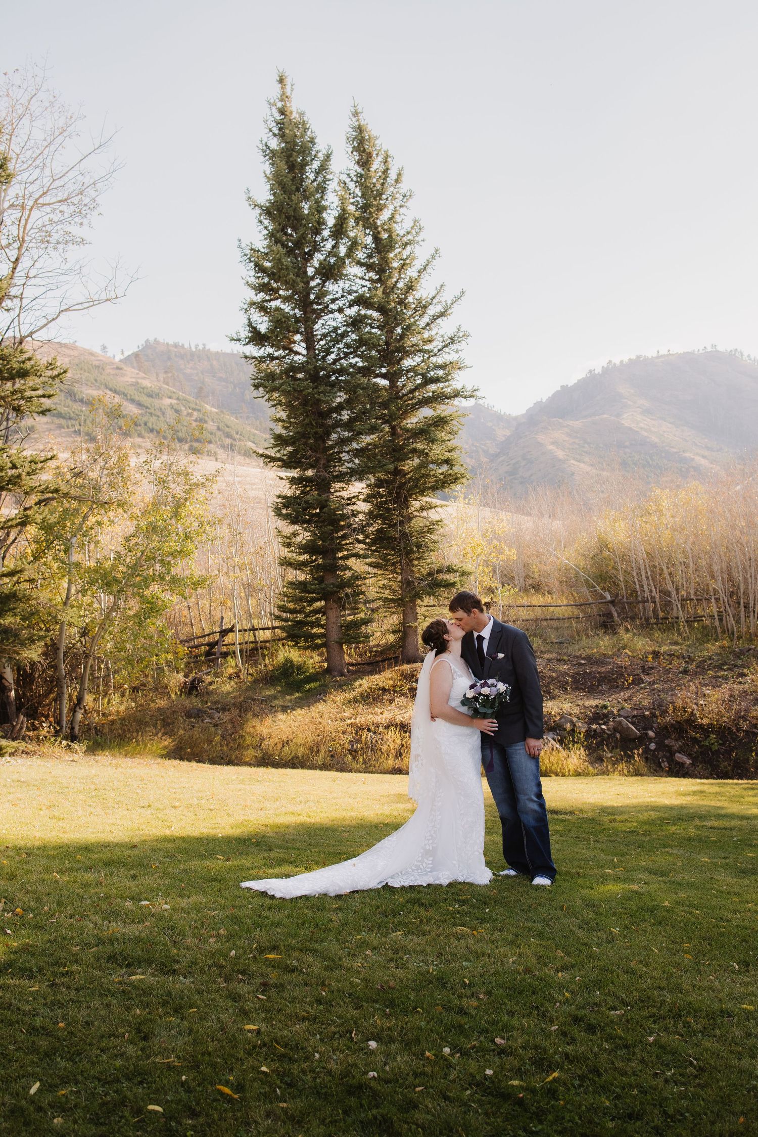 Bride and groom kiss on green lawn with tall pine trees and mountain backdrop. Wyoming wedding.