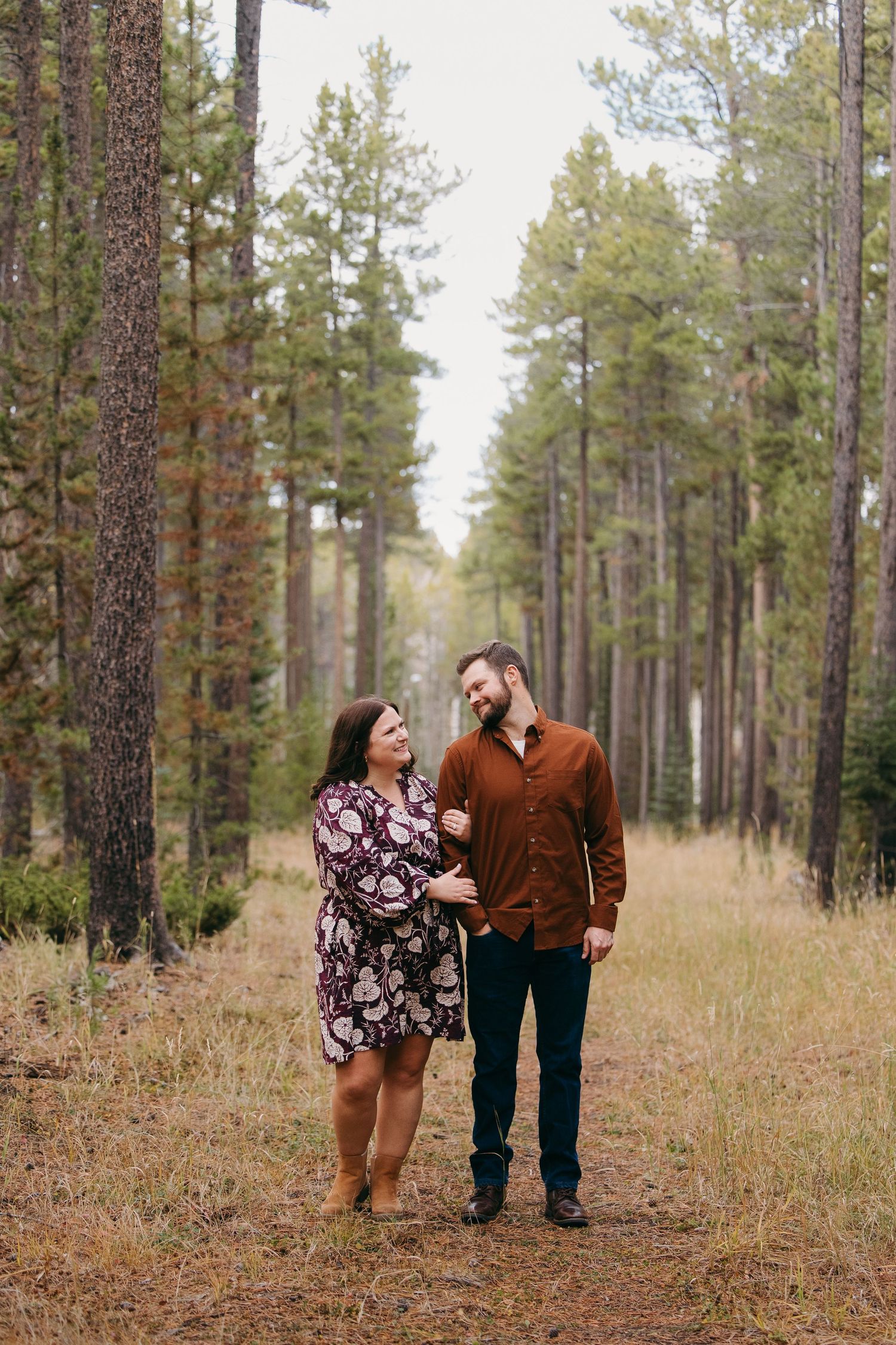 Couple in casual attire share a loving glance while walking through tall pine forest. Wyoming