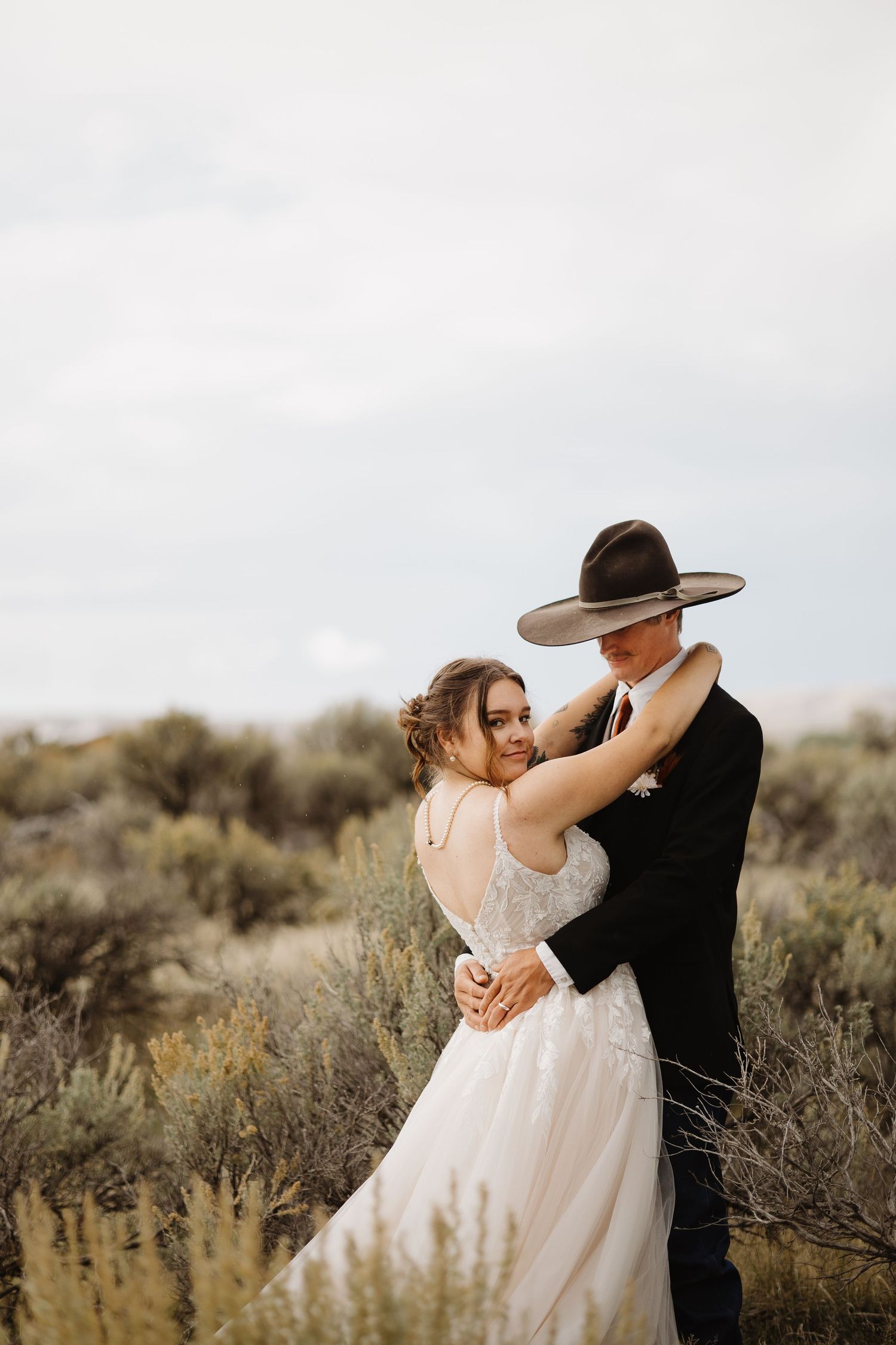 Bride and groom embrace in a desert landscape with sagebrush, groom wearing a wide-brim hat.