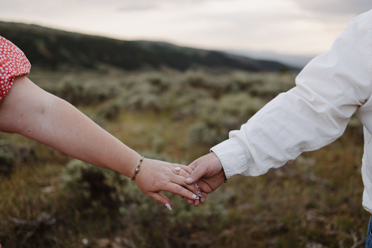 Close-up of couple holding hands in field with sagebrush landscape in soft background.
