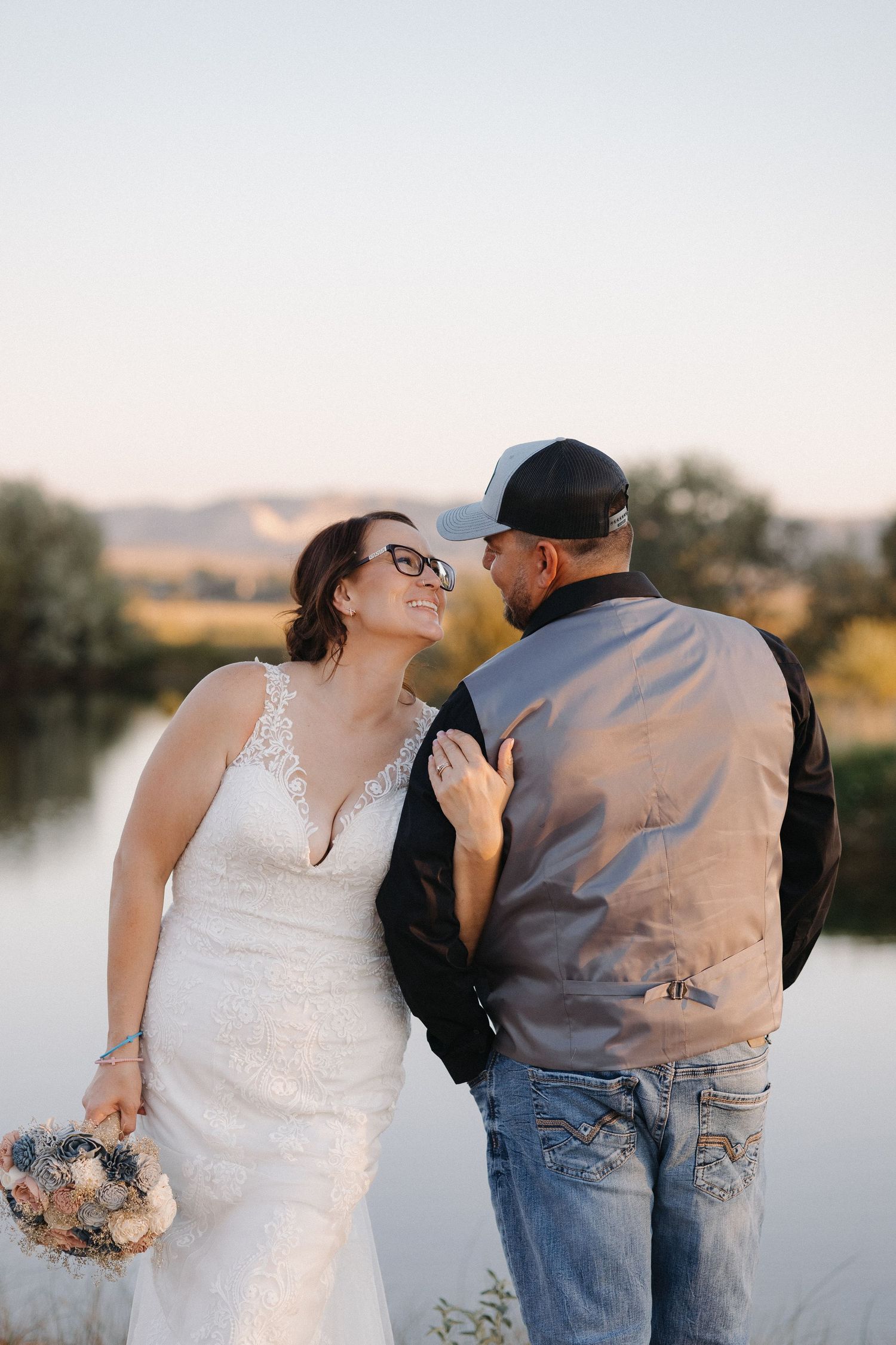 Bride in lace gown and groom share a romantic moment by a scenic lake at sunset.