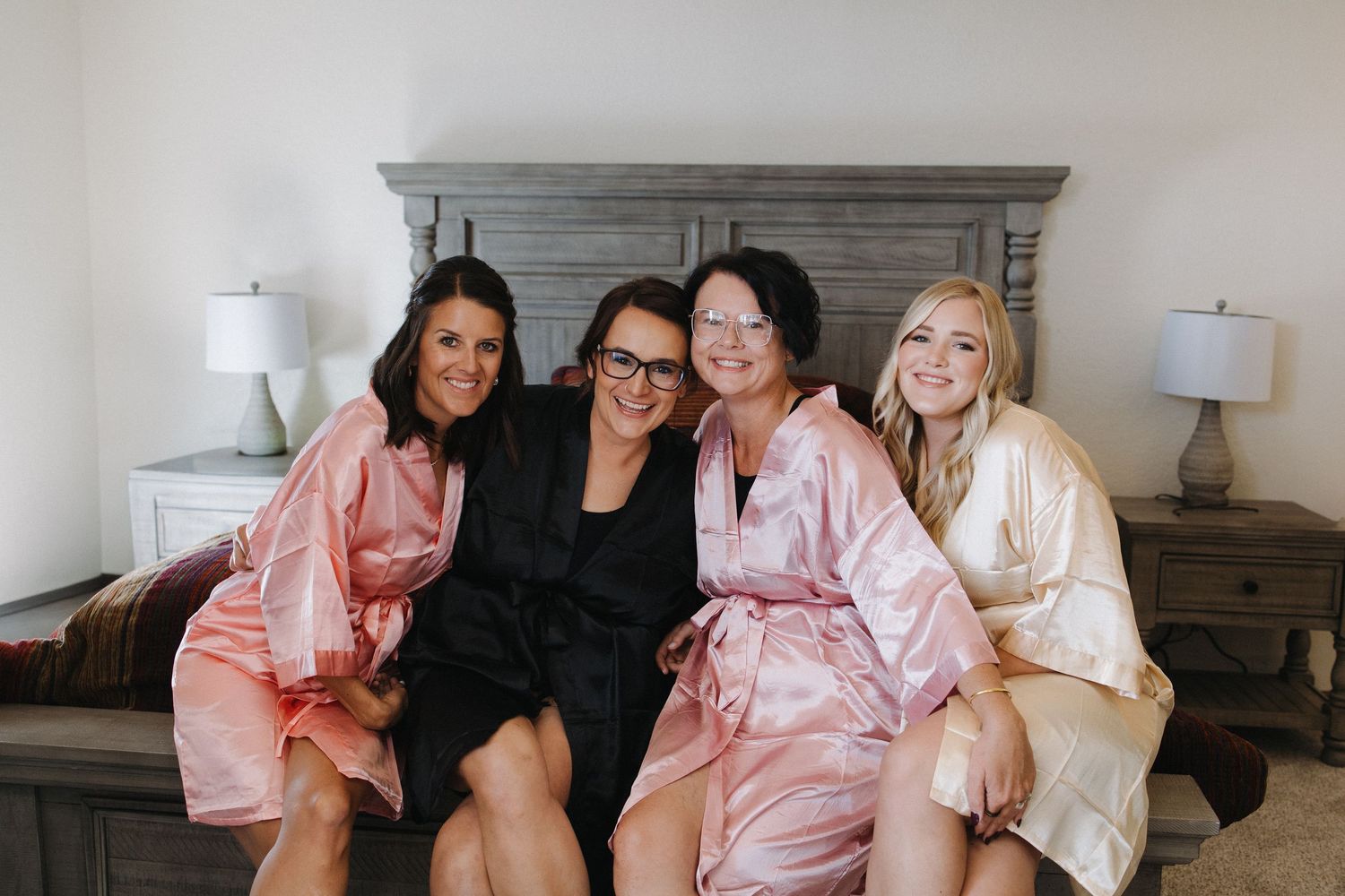 Four women in satin robes smile together on a bed during wedding preparations.