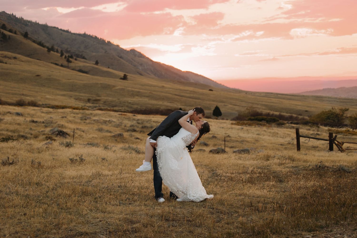 Groom dips bride in white gown at golden sunset over rolling Montana mountain meadow landscape.Wyoming wedding.