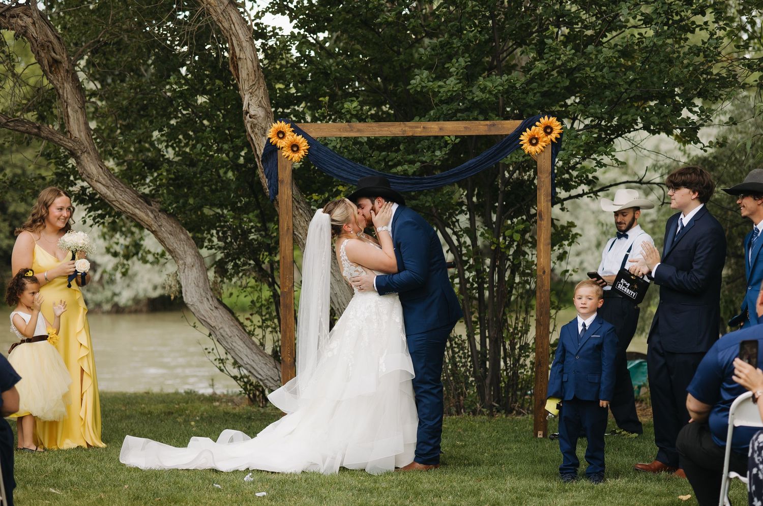 Bride and groom share their first kiss under a sunflower-adorned wooden arch at an outdoor wedding.