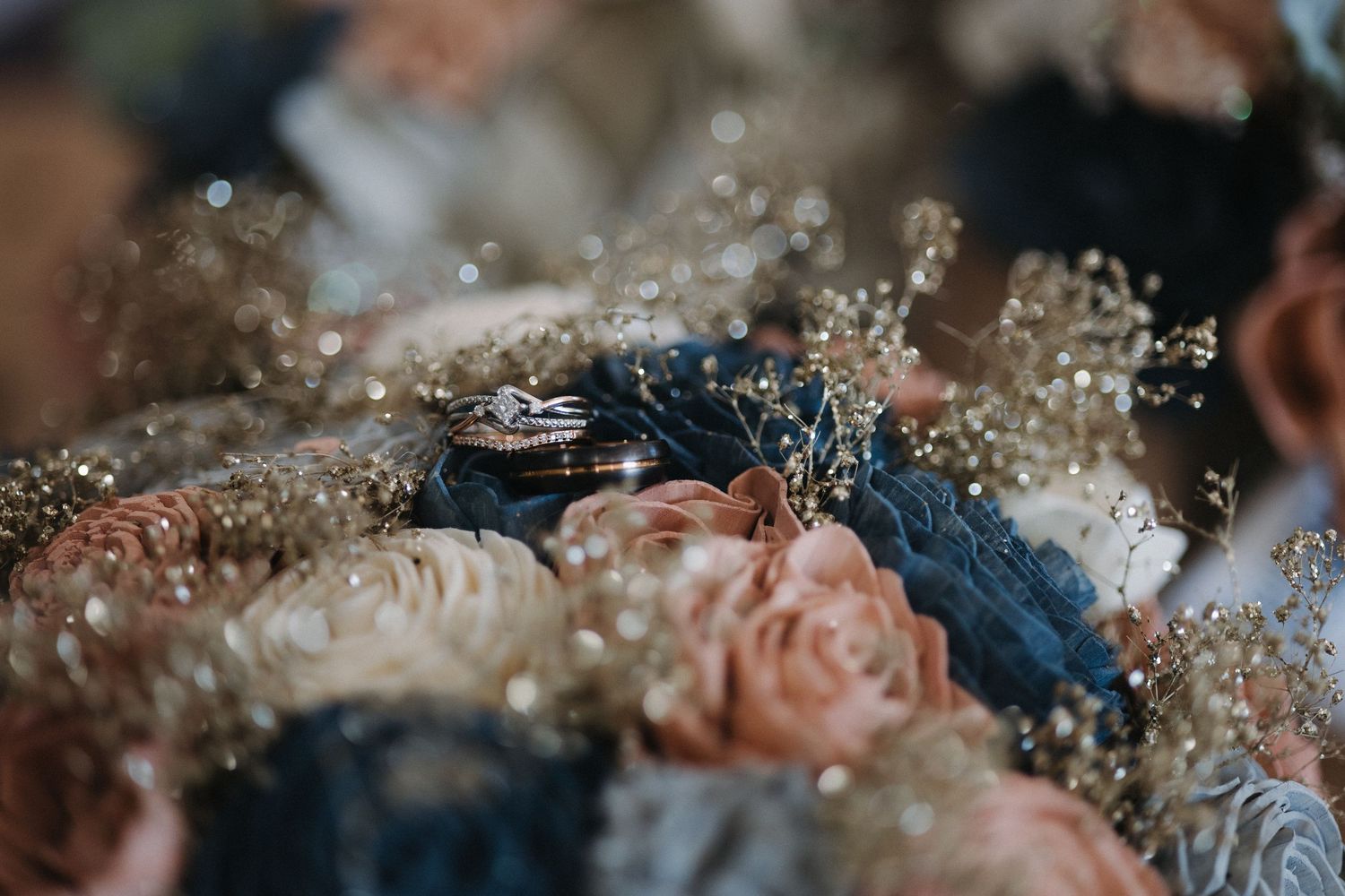 Close-up of wedding rings nestled among navy blue and blush fabric flowers with gold beaded accents.