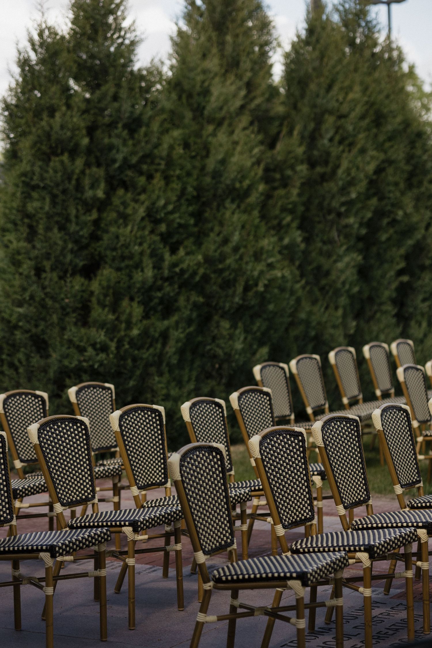 Rows of black and gold wicker chairs set up outdoors for a wedding ceremony near evergreen trees.