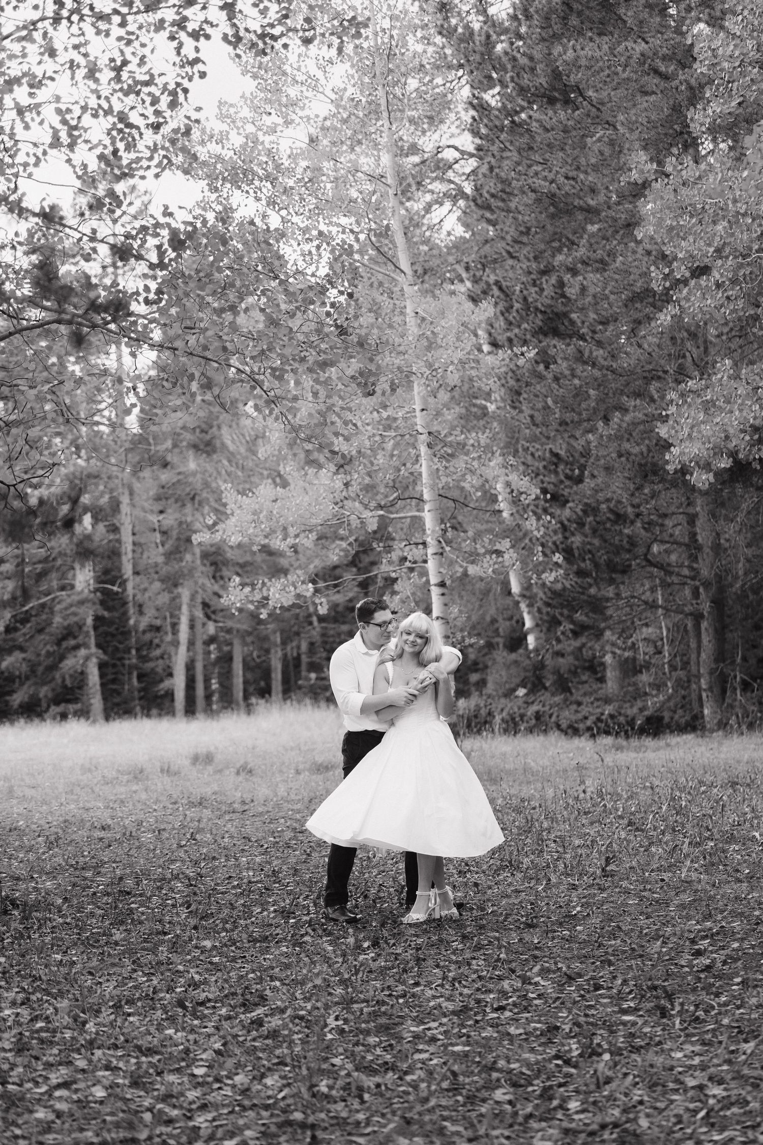 Black and white photo of bride and groom dancing together among tall trees in autumn forest. Wyoming wedding.