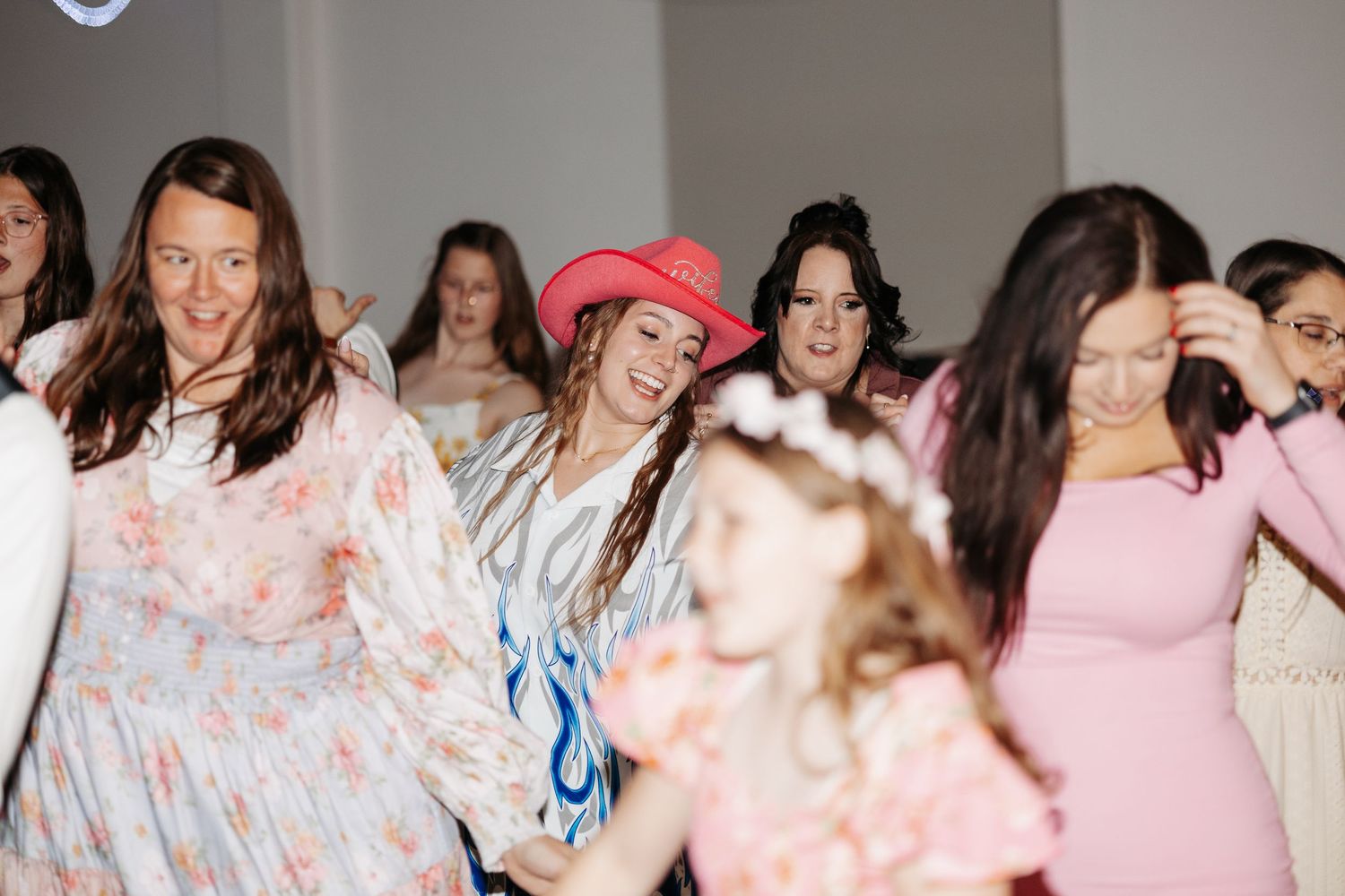 Guests dance joyfully at a wedding reception, with one woman wearing a pink cowboy hat.