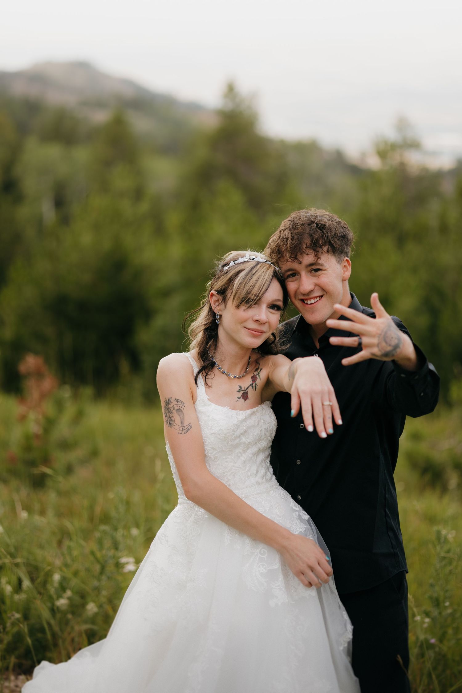 Young bride and groom posing together in a green meadow, groom showing off wedding ring.