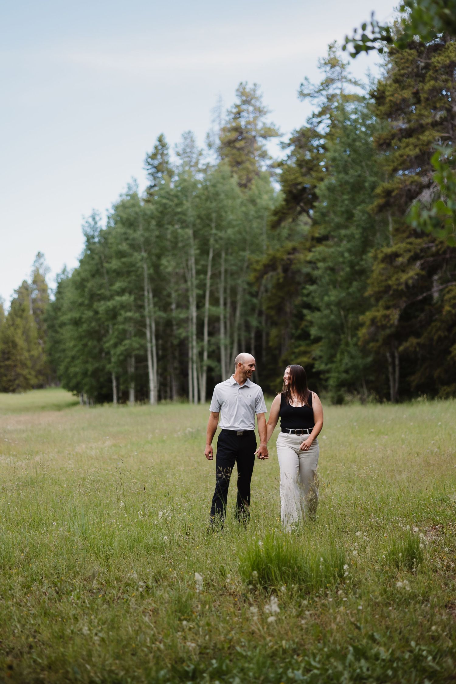 Couple holding hands walking through a lush green meadow surrounded by tall pine trees.
