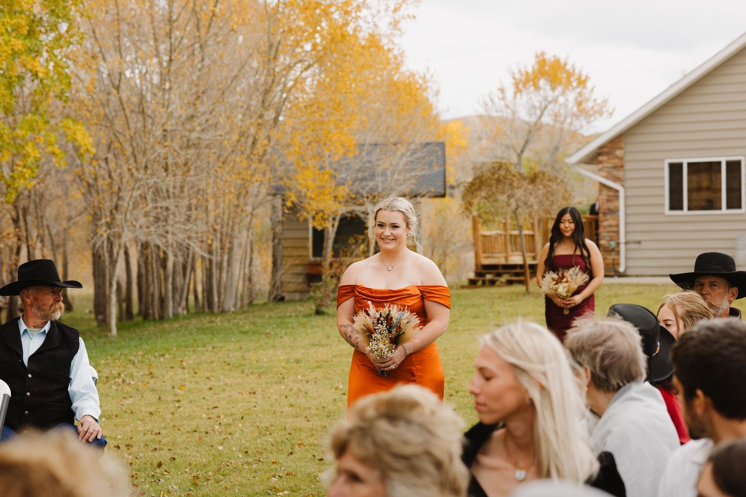 Bridesmaid in orange off-shoulder dress walks down the aisle during outdoor fall wedding.