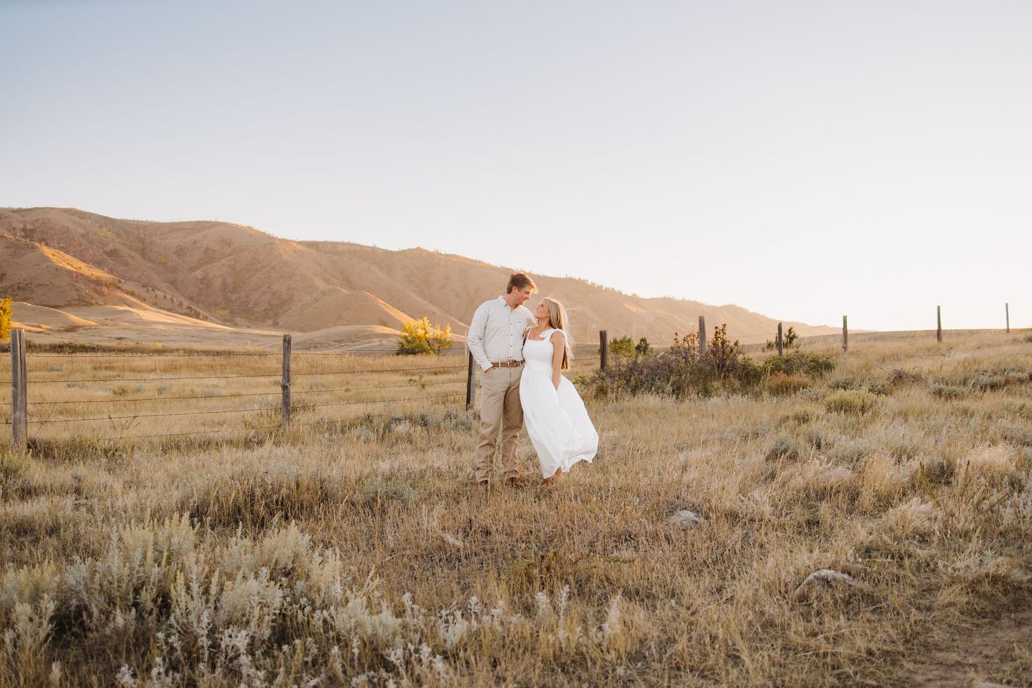 Bride and groom share a romantic moment in a sun-drenched open field with rolling hills behind them.