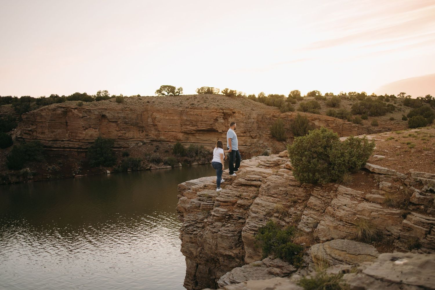 Couple standing hand in hand on rocky cliffs overlooking a calm river at dusk in a canyon landscape.