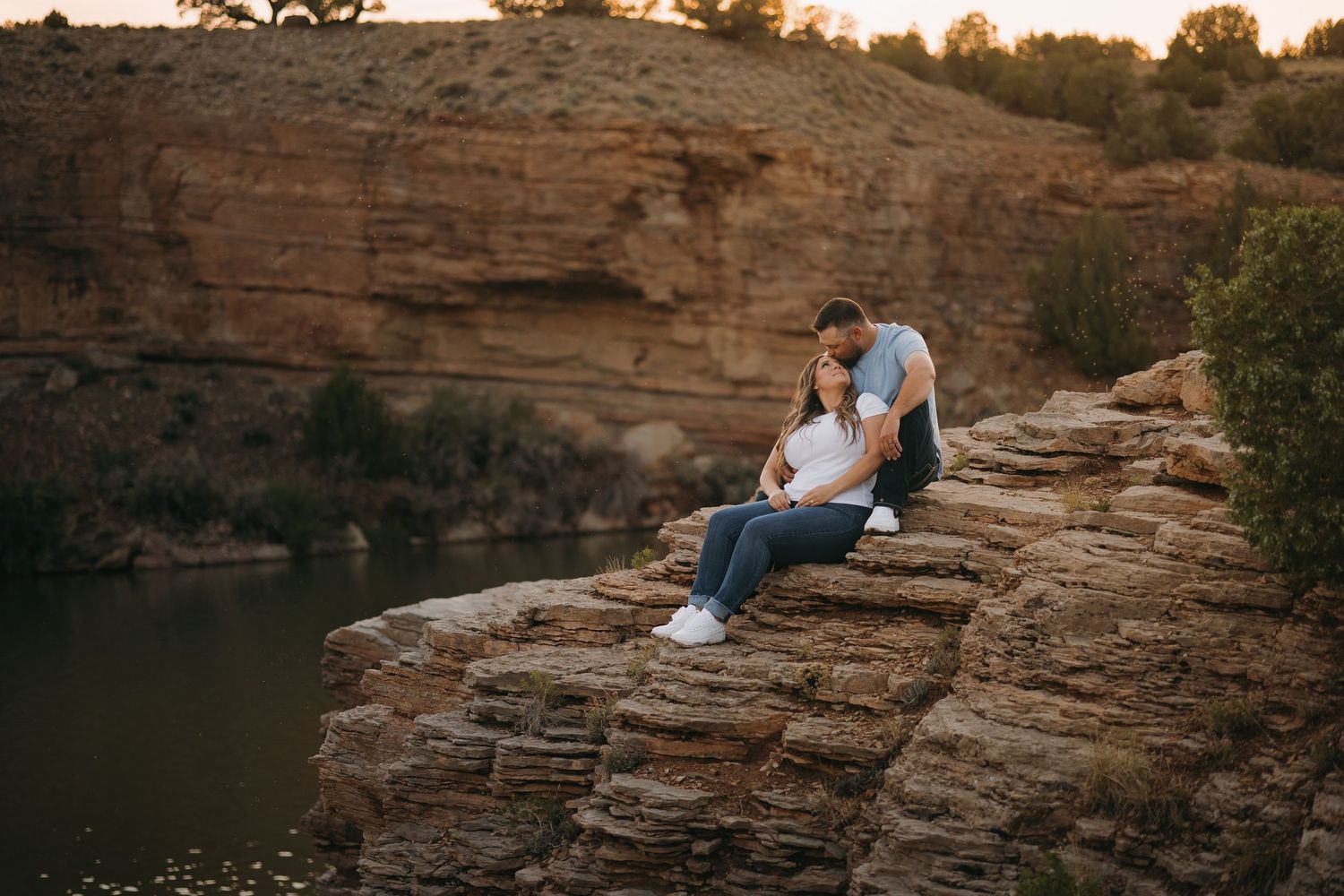 Couple sits closely together on rocky cliffs overlooking a river at warm golden sunset.