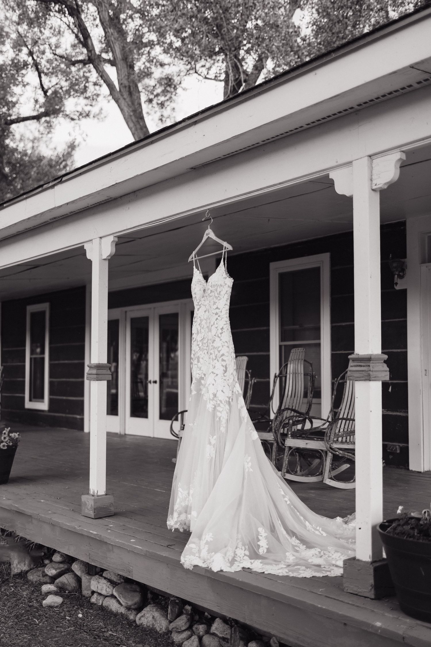 Elegant lace wedding gown hangs on a porch of a rustic farmhouse in black and white photo.