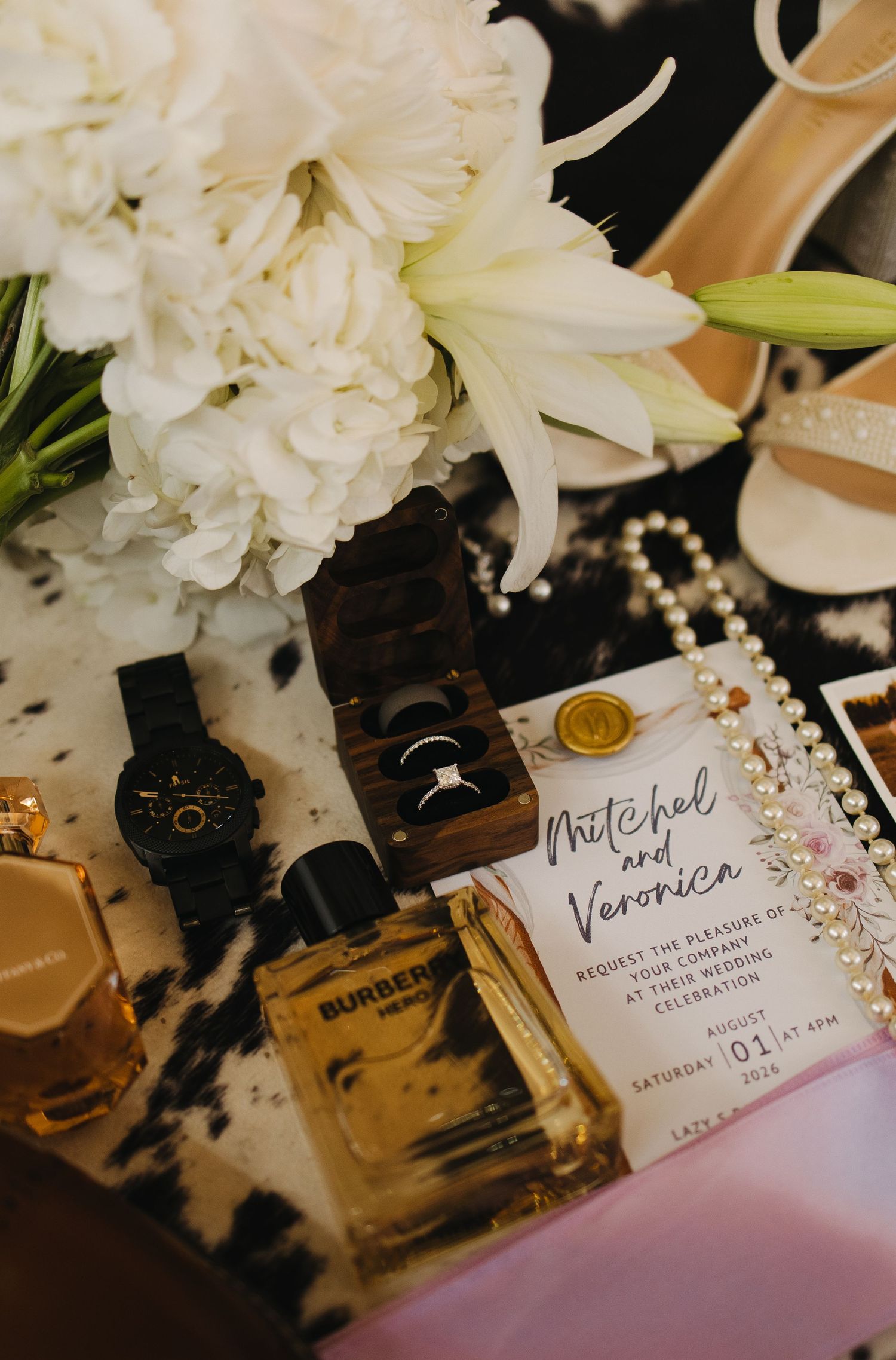 Wedding flat lay featuring invitation for Mitchel and Veronica, rings, Burberry cologne, watch, and white flowers.