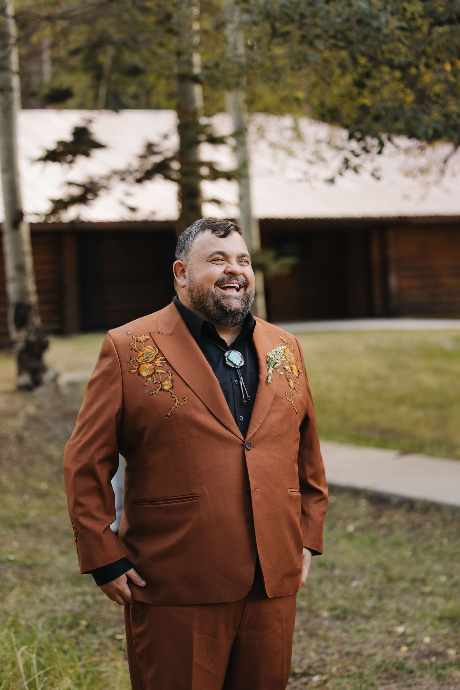 Smiling groom in rust brown embroidered western suit with bolo tie stands outside log cabin venue. Wyoming wedding.