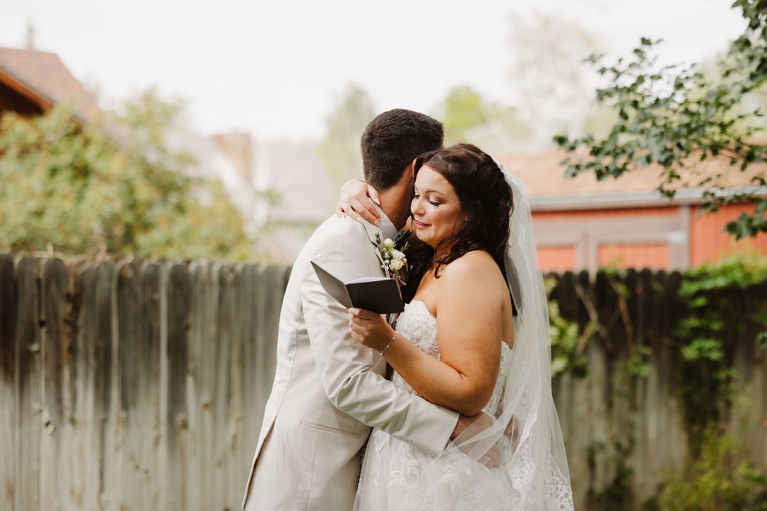 Bride in strapless lace gown embraces groom in light suit while smiling near a wooden fence.