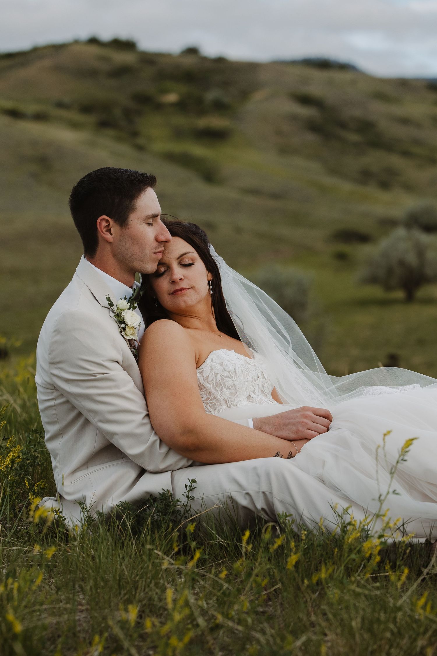 Bride and groom sit peacefully in a green hillside meadow, sharing a tender embrace at dusk.