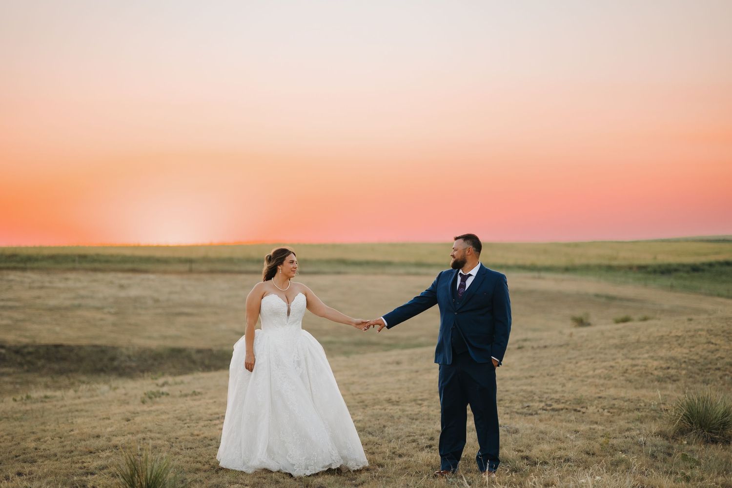 Bride in ball gown and groom in navy suit holding hands in a golden field at sunset.