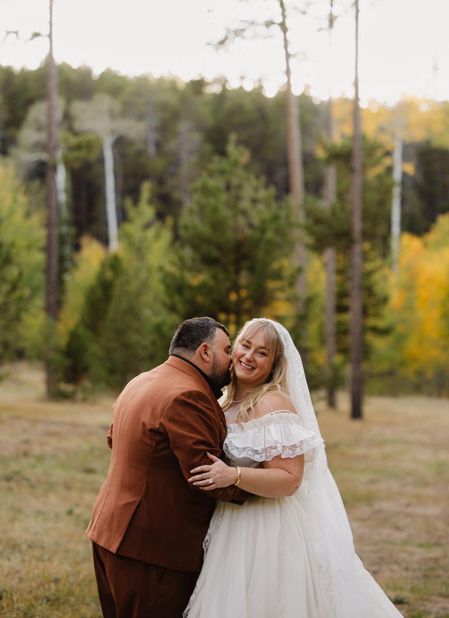 Bride in off-shoulder white gown laughs as groom in brown suit kisses her cheek in autumn forest. Wyoming wedding.