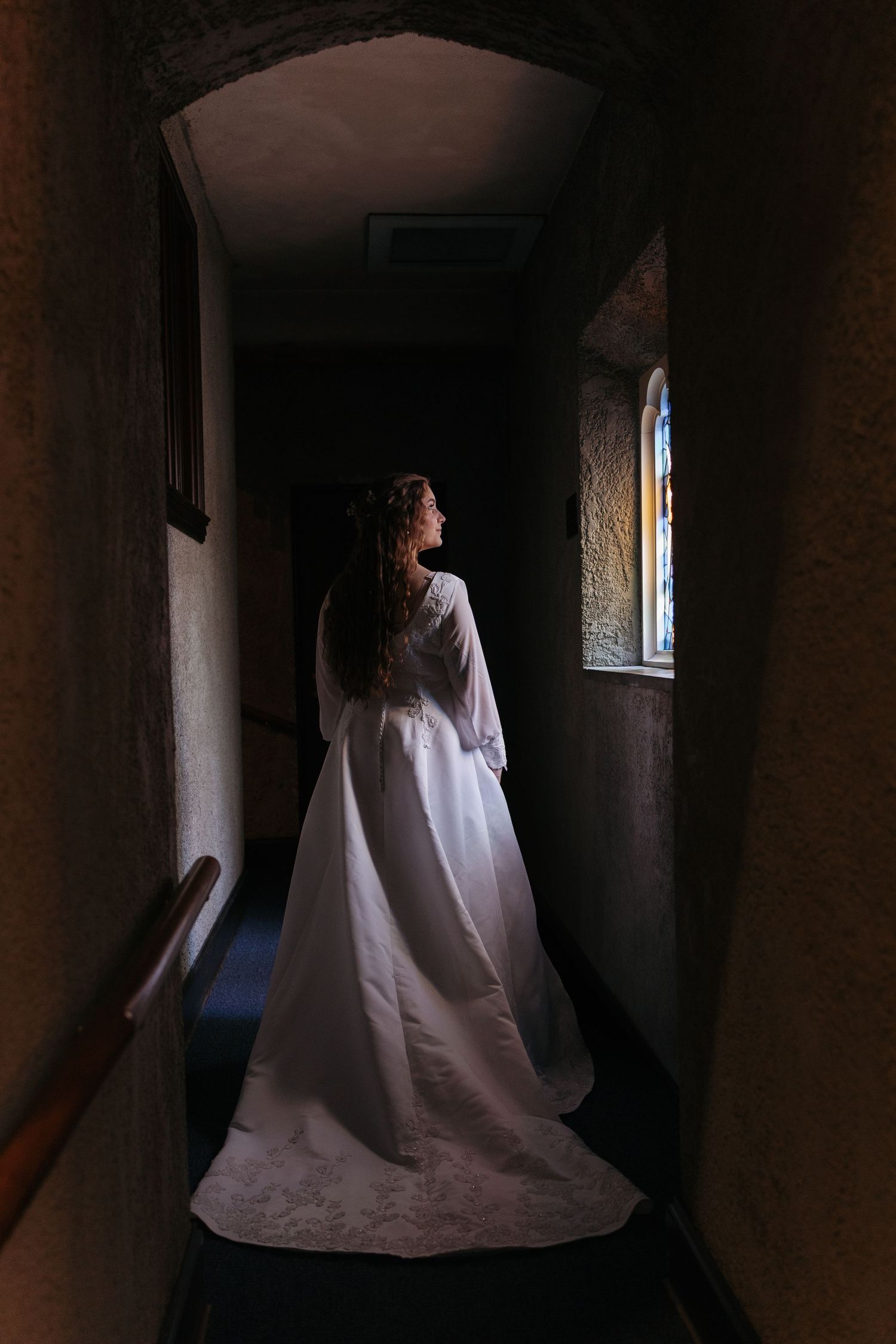 Bride in elegant white wedding gown standing in a dimly lit church hallway near a stained glass window.