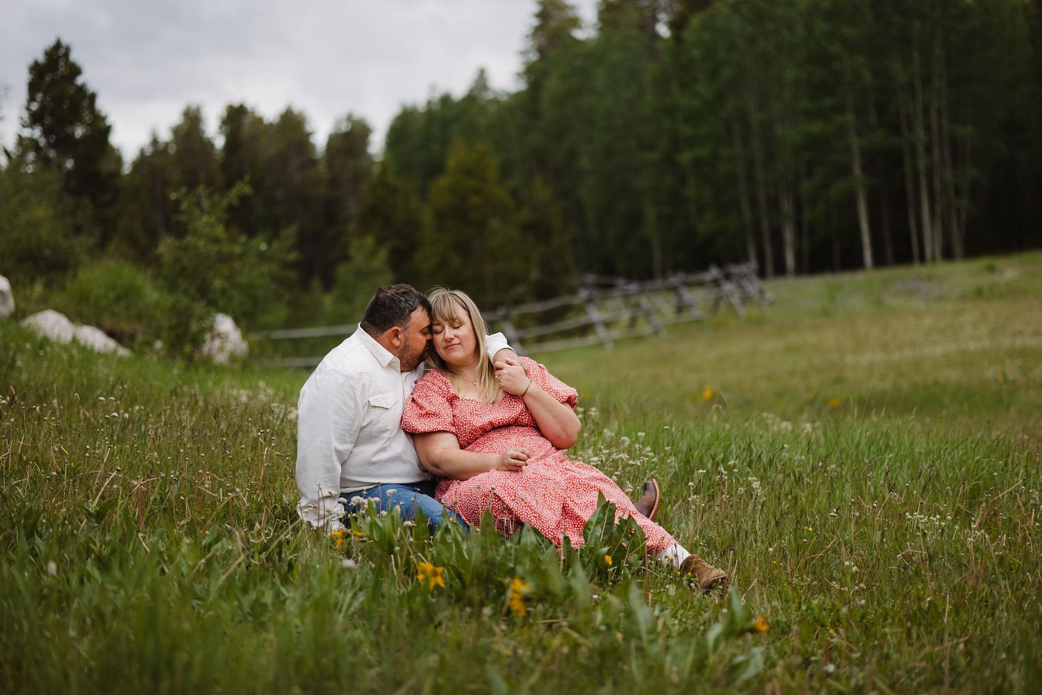 Couple sitting in a lush green meadow surrounded by trees during an outdoor engagement photo session.