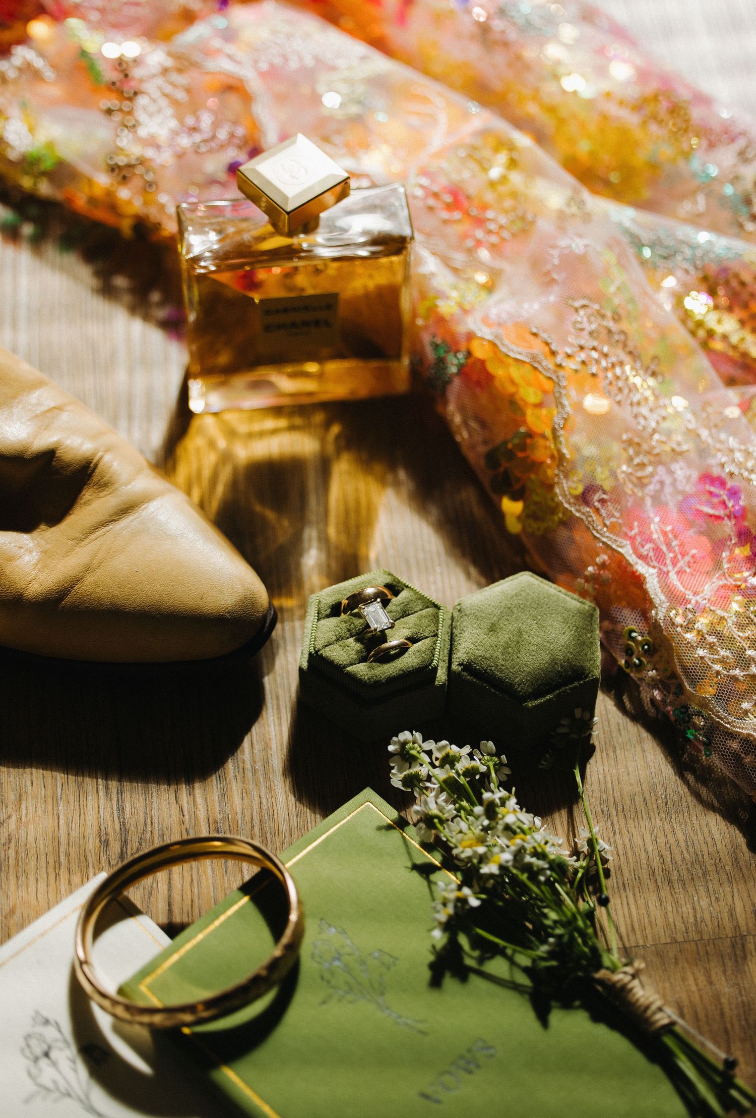 Elegant wedding flat lay with Chanel perfume, gold ring, green velvet ring box, and colorful embroidered fabric.