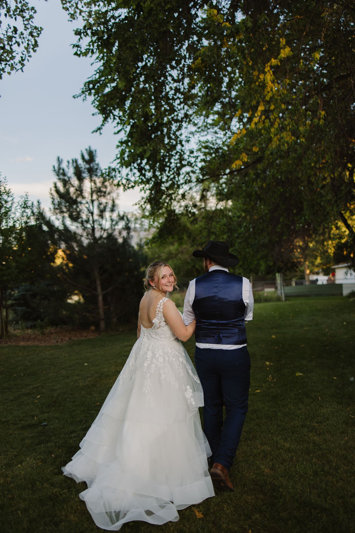 Smiling bride in lace gown glances back while walking with groom in navy vest through garden.