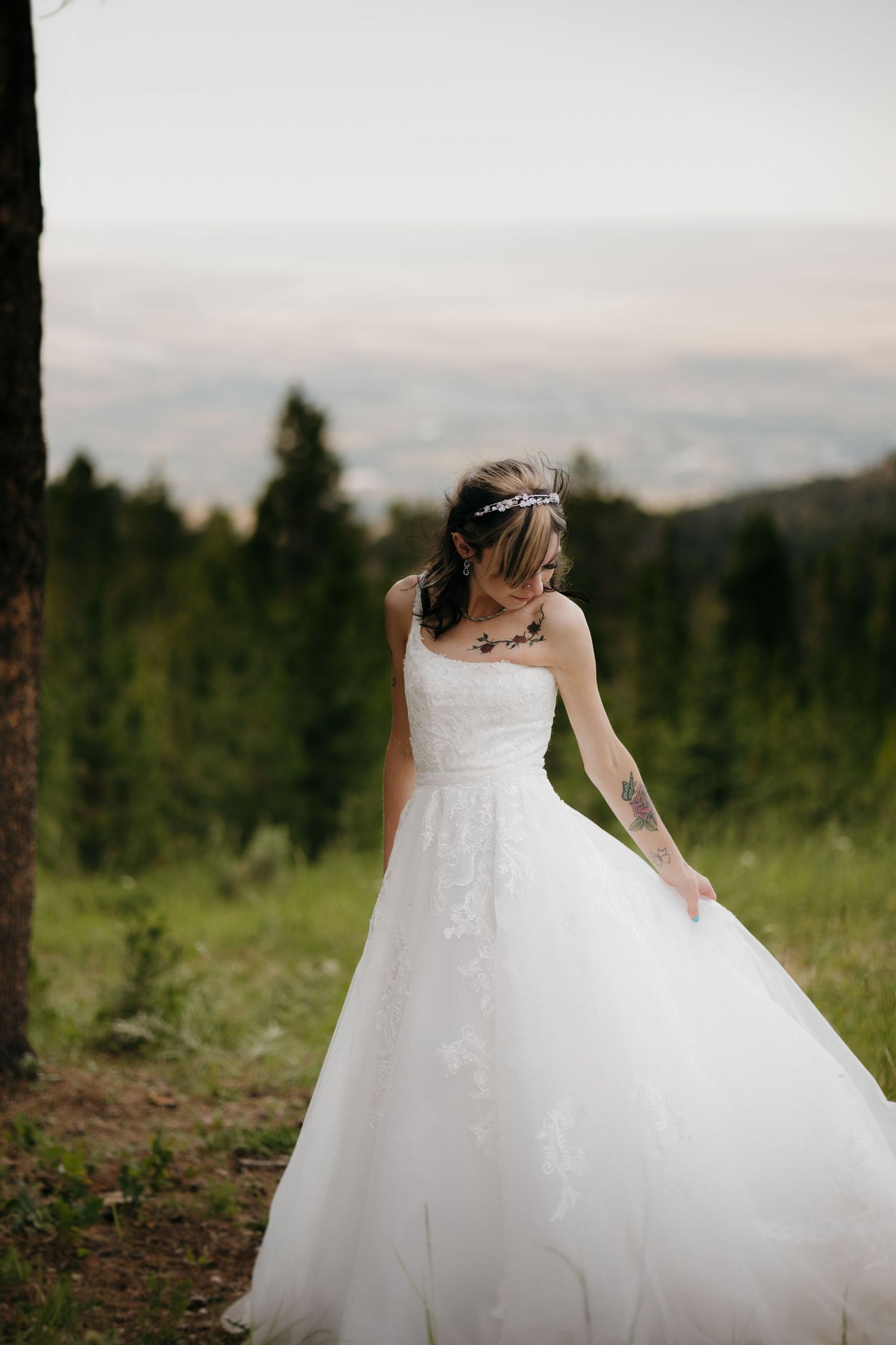 Bride in white lace ball gown with tattoo and crystal headpiece posing in a mountain forest.