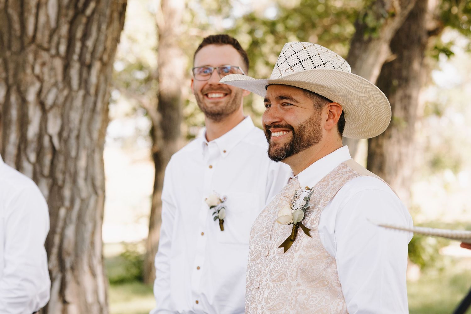 Smiling groom in cowboy hat and vest stands at the altar with his best man beside him outdoors.
