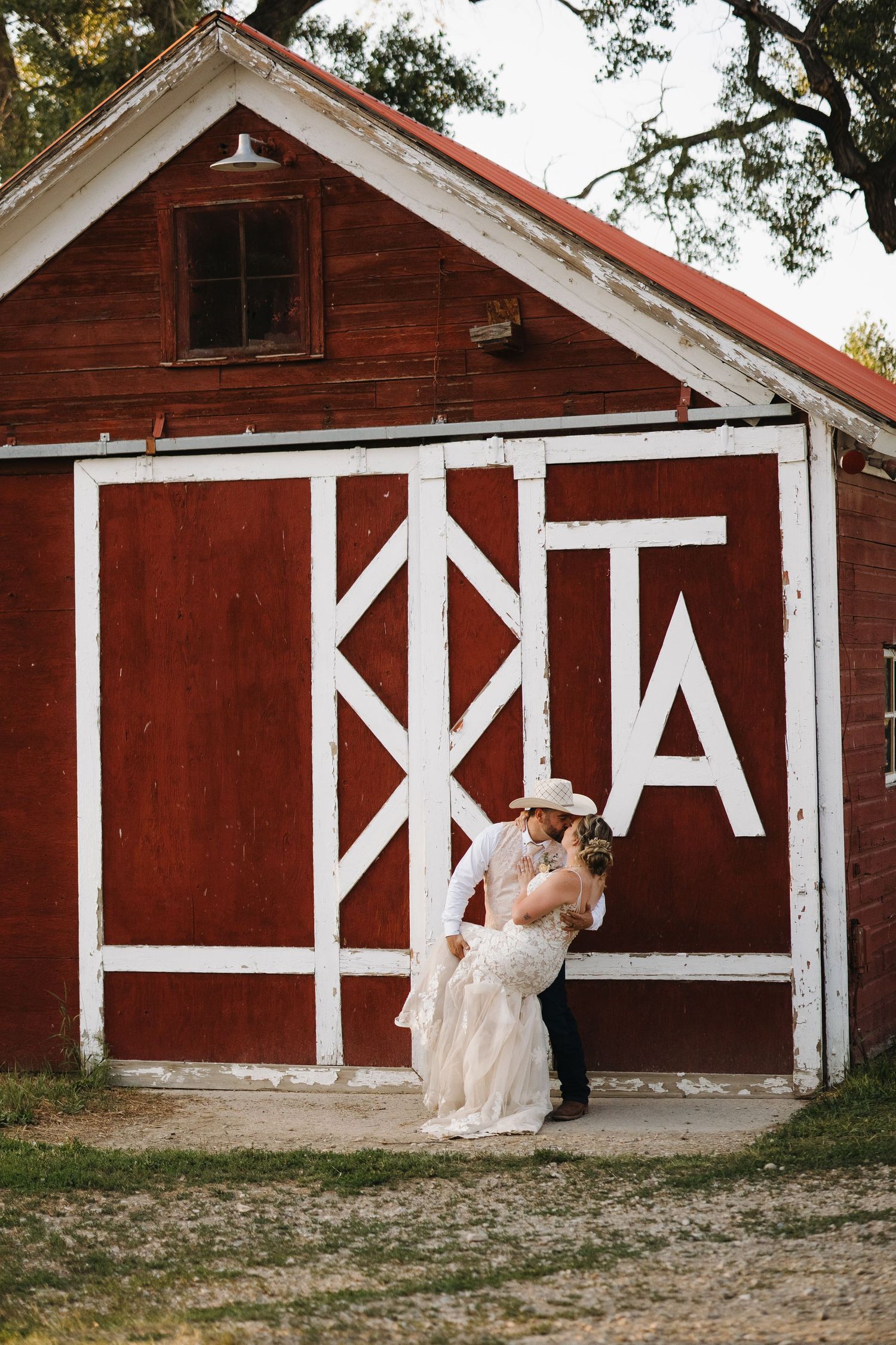 Bride and groom share a romantic kiss in front of a rustic red barn with white trim at a country wedding.