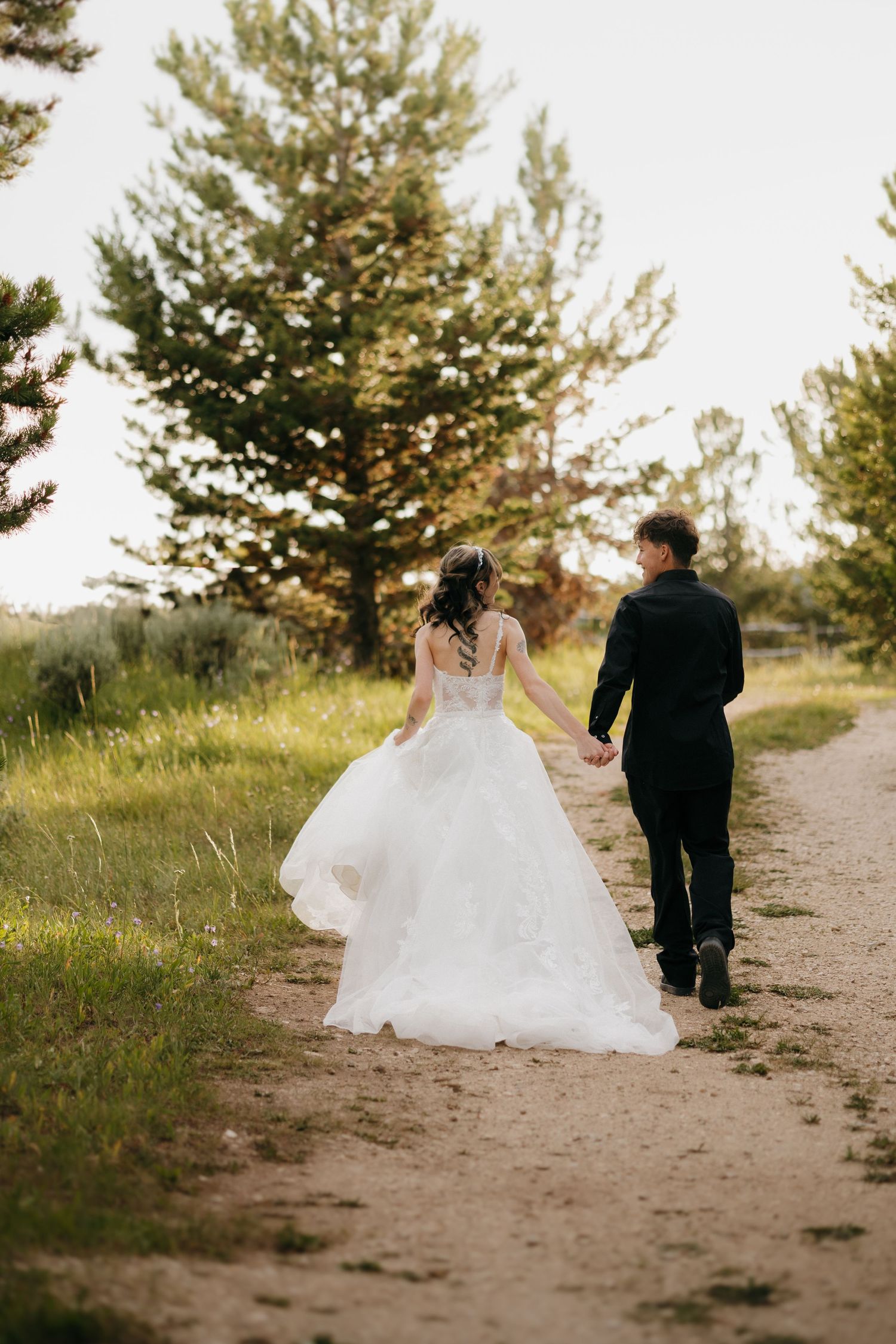Bride and groom walk hand-in-hand down a scenic dirt path surrounded by lush green trees at golden hour.