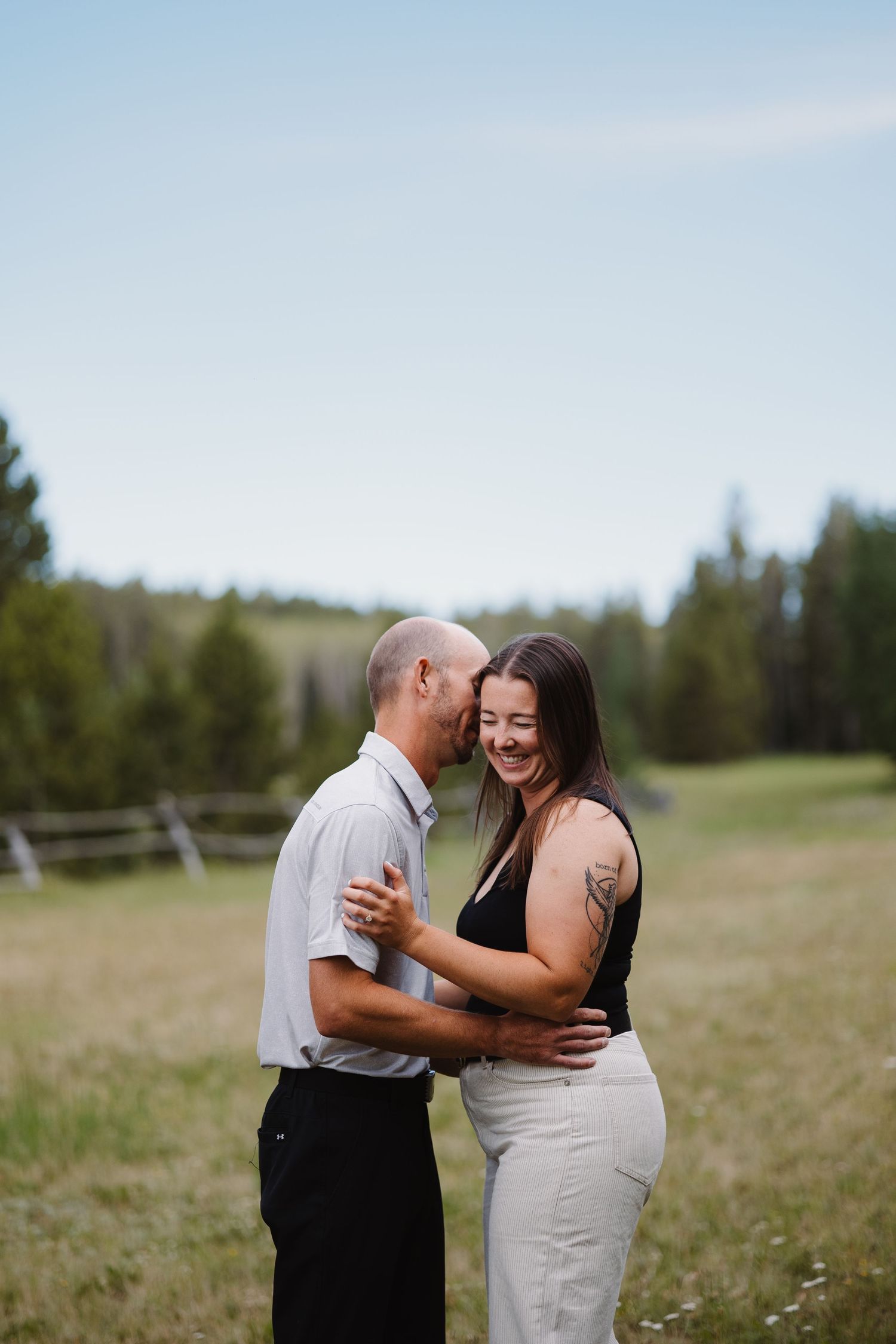 Happy couple laughing and embracing in an open field with pine trees in the background.