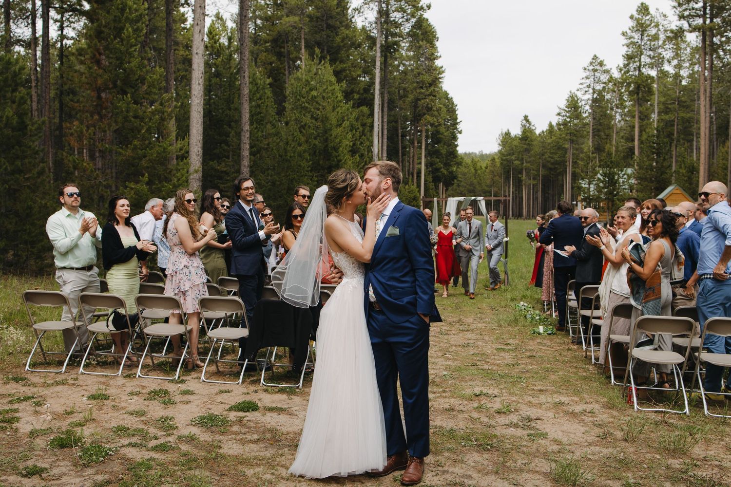 Bride and groom share first kiss at outdoor forest wedding ceremony surrounded by cheering guests.