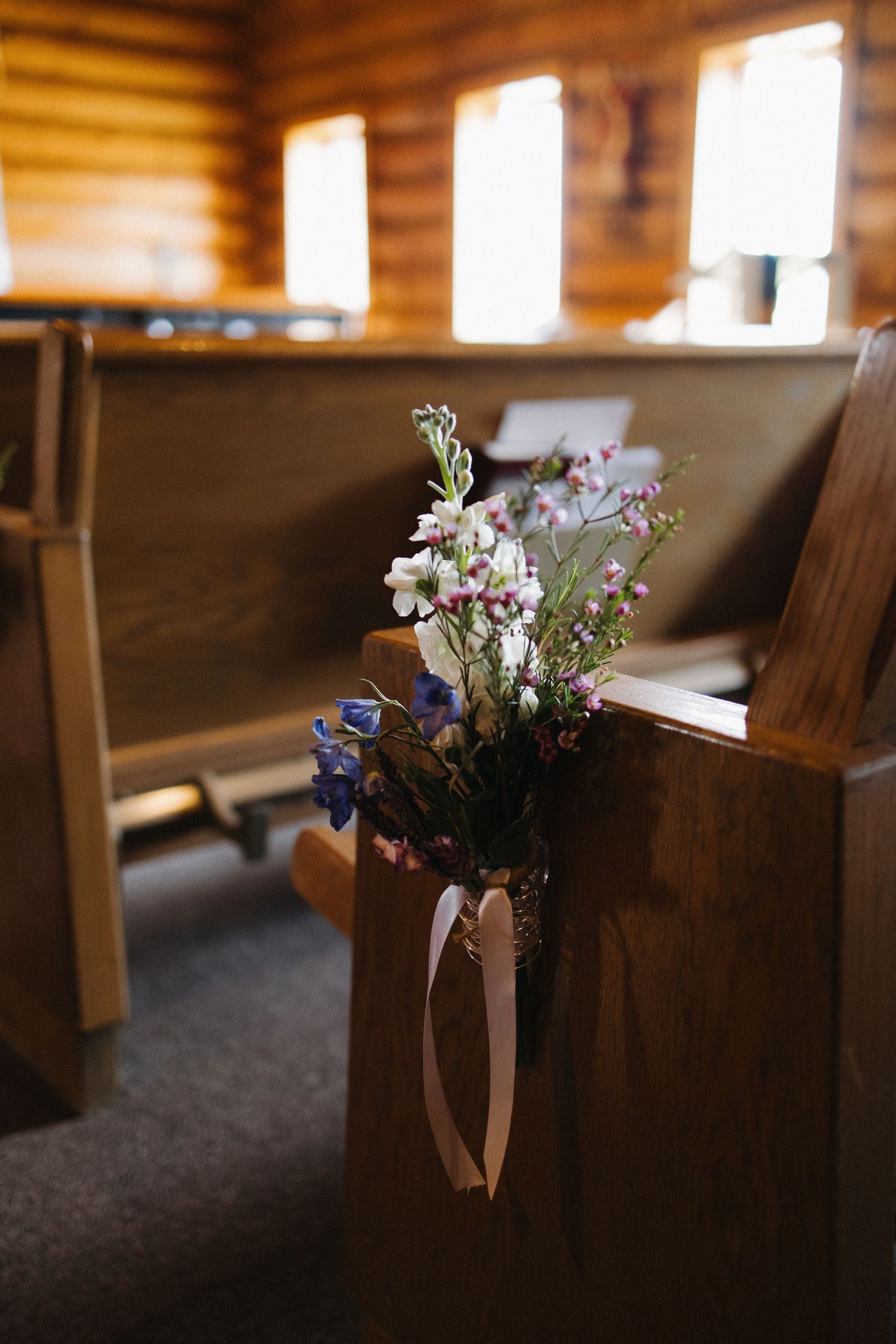 Wildflower wedding pew decoration with pink, white, and blue blooms tied with ribbon in a log chapel.