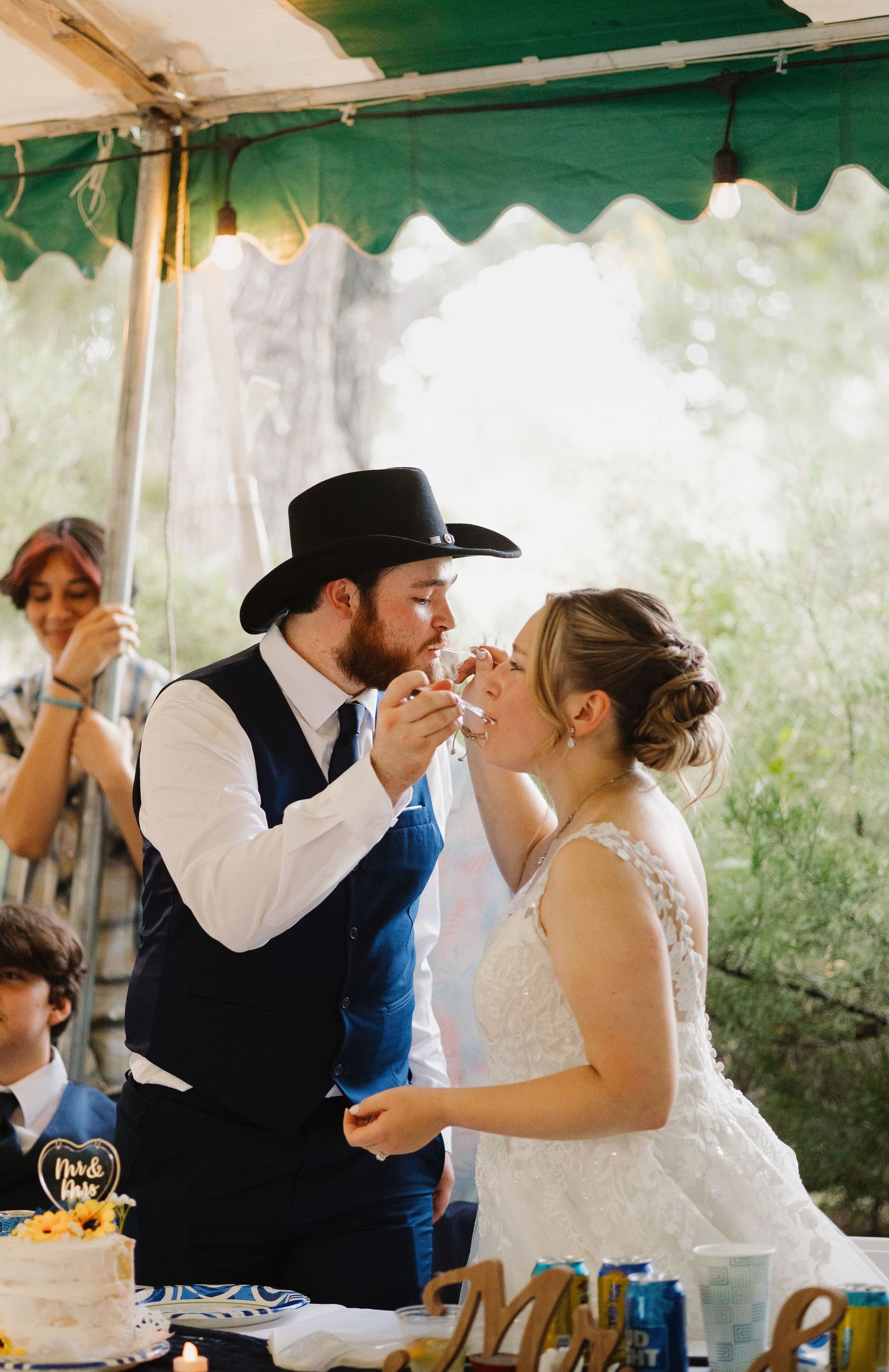 Groom in a cowboy hat feeds wedding cake to his bride at an outdoor rustic wedding reception.