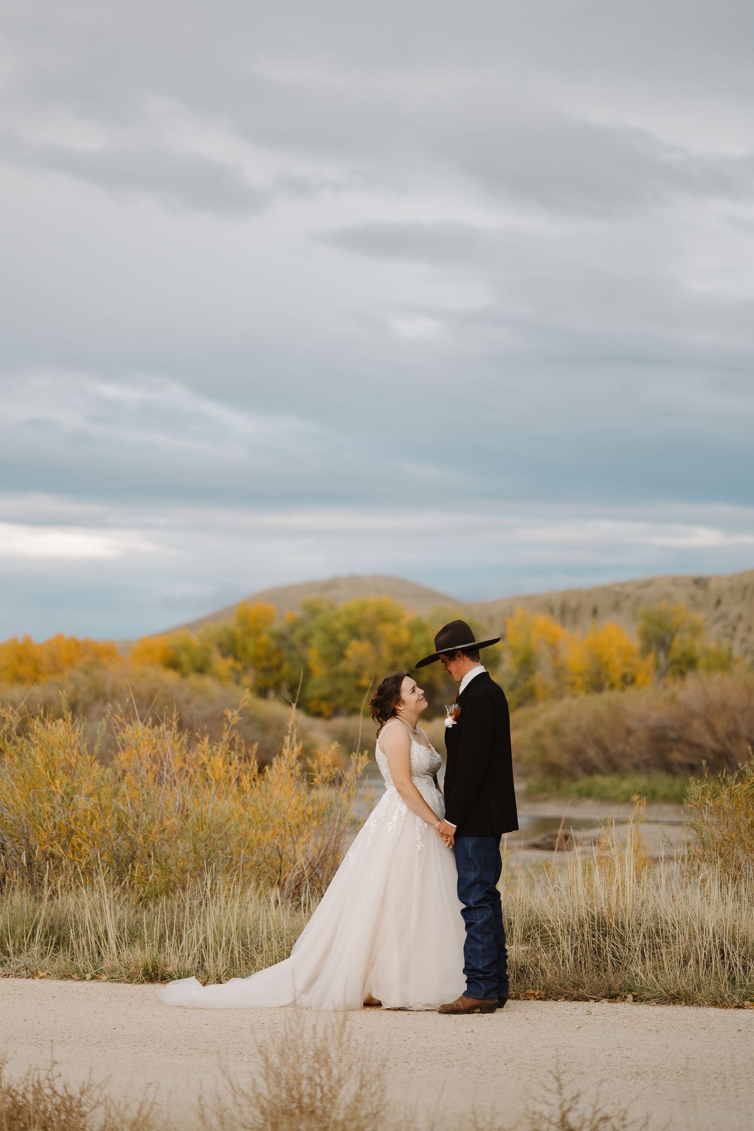 Bride and groom hold hands on a dirt path surrounded by golden fall foliage.